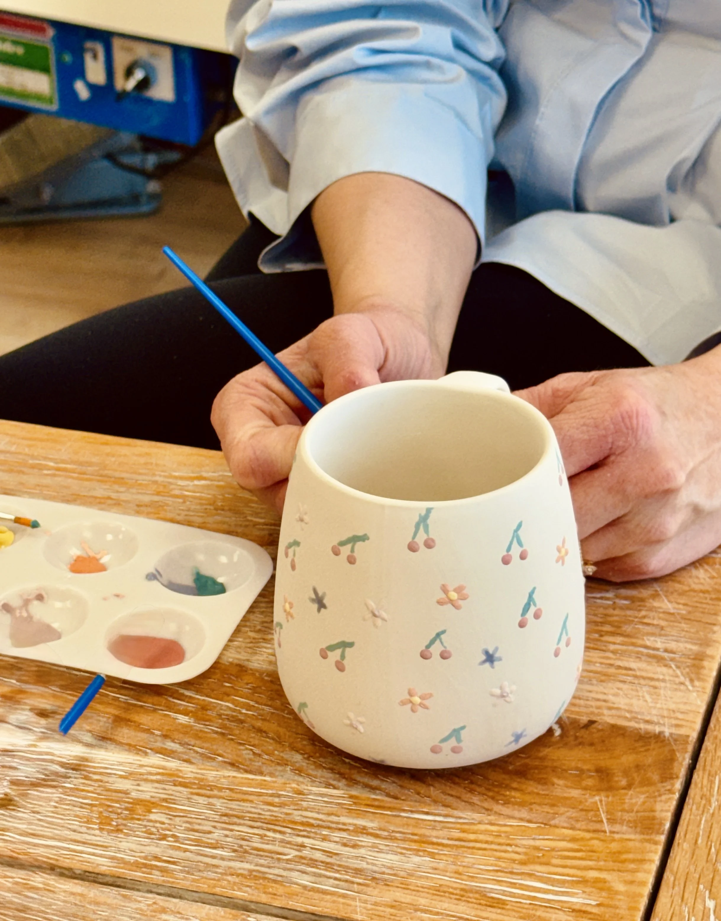 Une personne peint une tasse blanche décorée de petites cerises et de fleurs avec une peinture bleue, utilisant une palette de peinture avec différentes couleurs.