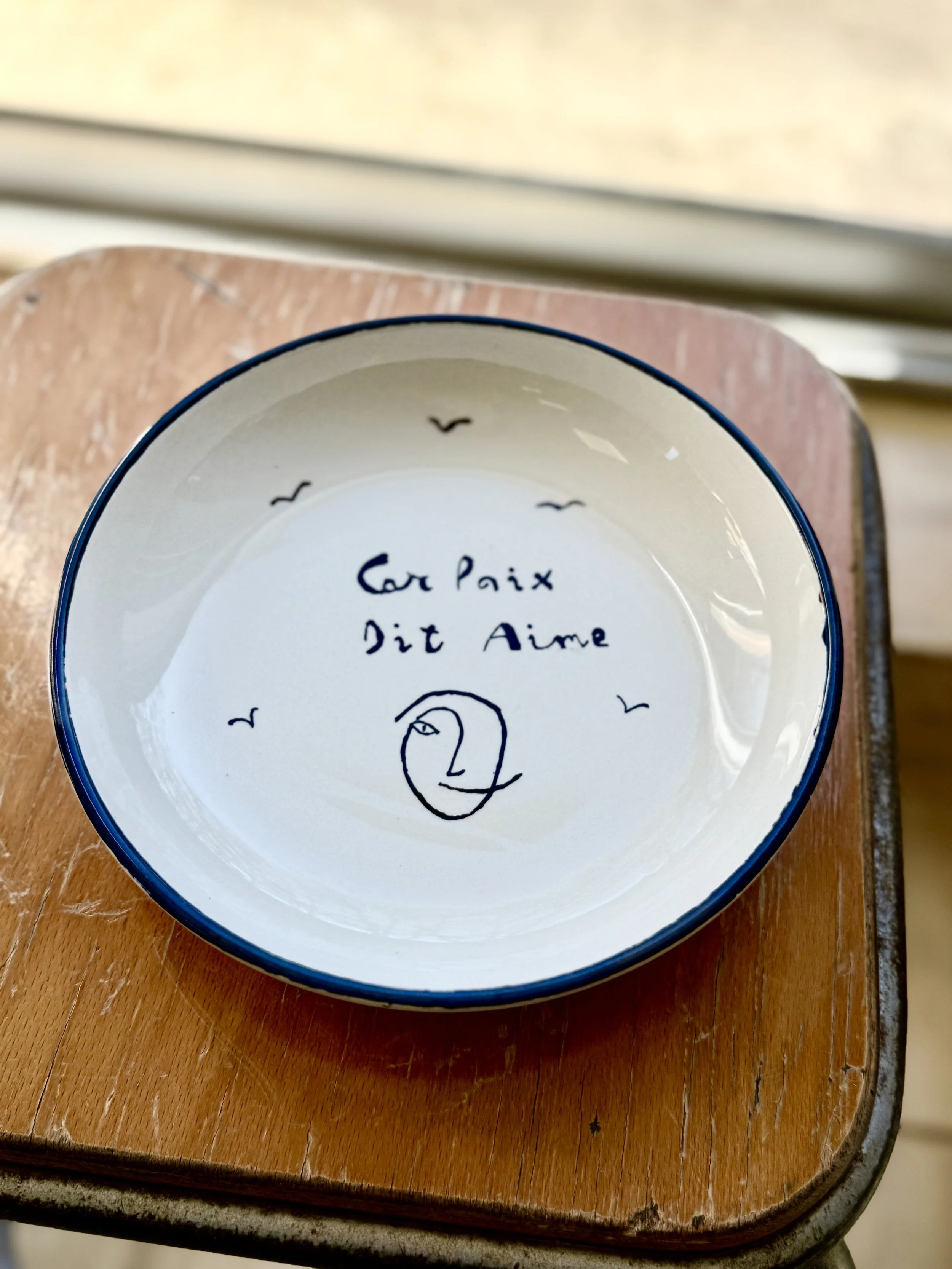 Un bol blanc avec une inscription manuscrite en français et un dessin, placé sur une petite table en bois. La phrase dans le bol dit: "Car Paix Dit Aime" et il y a un dessin d'un visage stylisé. Autour, des petits oiseaux sont dessinés.