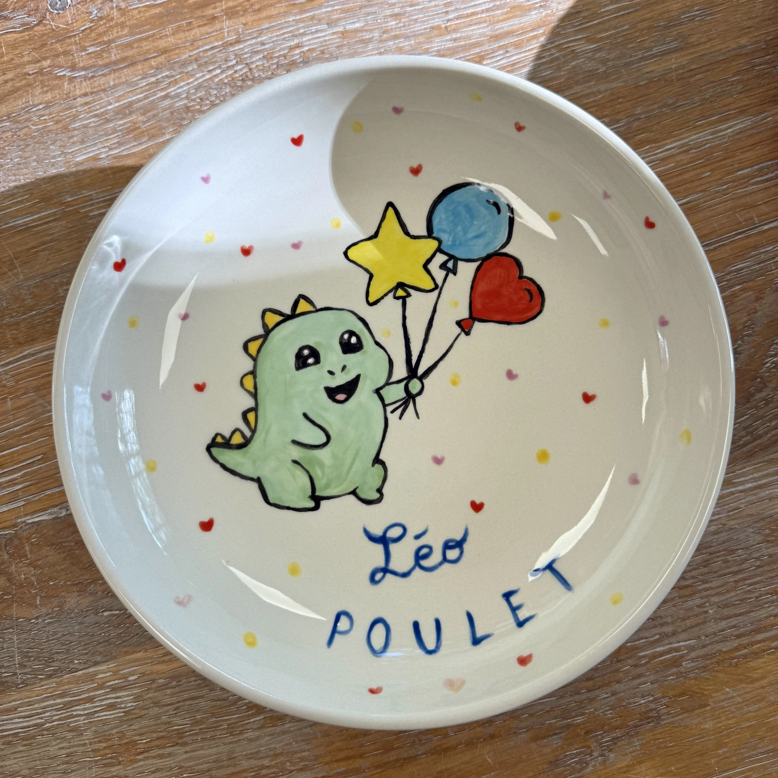 Assiette décorée avec un dessin d'un dino vert tenant trois ballons en forme de cœur, une étoile et un cercle, avec le nom «Léo Poulet» écrit en bleu, entourée de petits cœurs et points colorés.