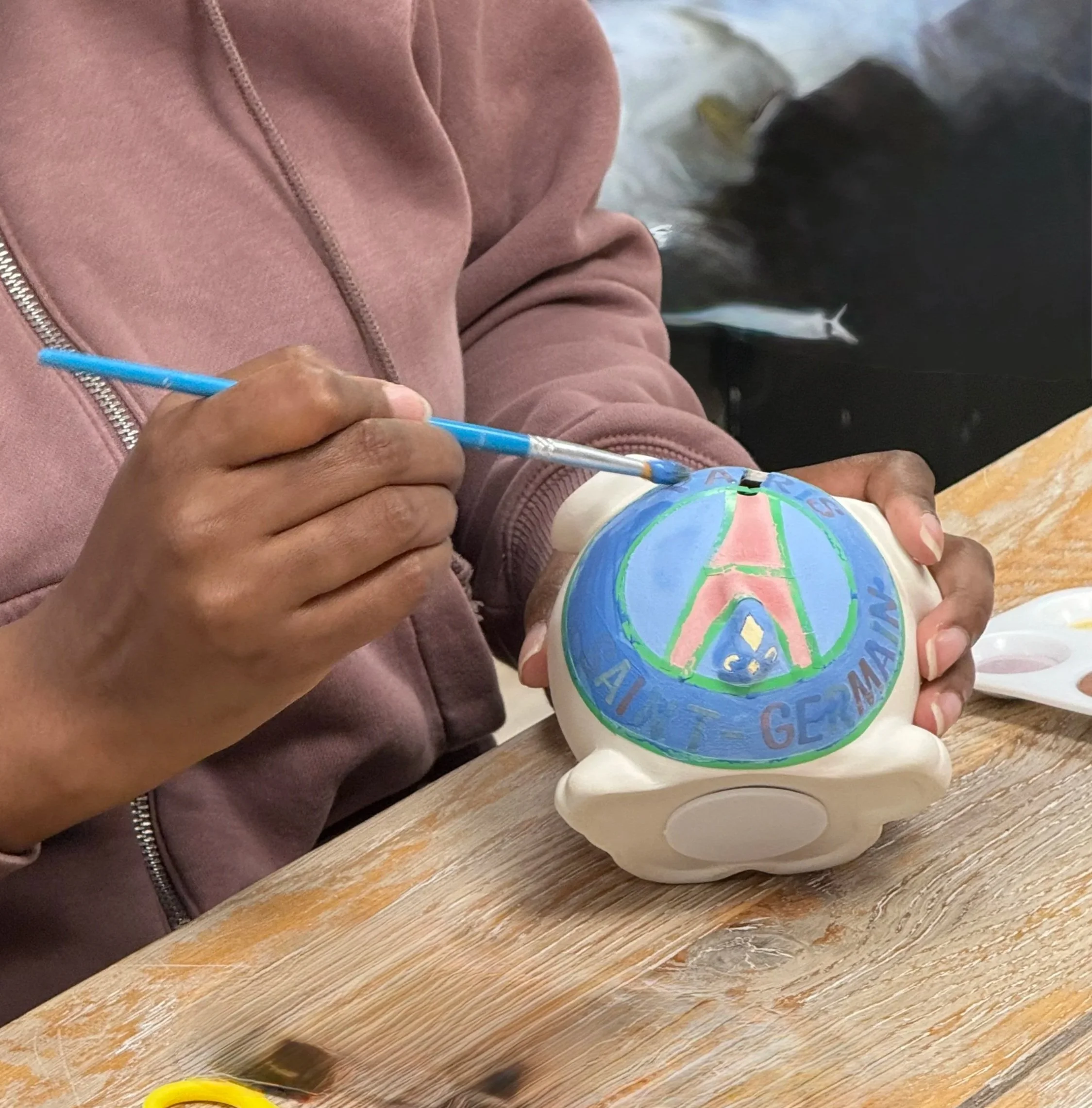 Personne en train de peindre un objet en céramique avec un pinceau bleu. L'objet est de forme ronde et beige, avec un dessin coloré de style graffiti représentant une tour Eiffel et des mots en français autour.