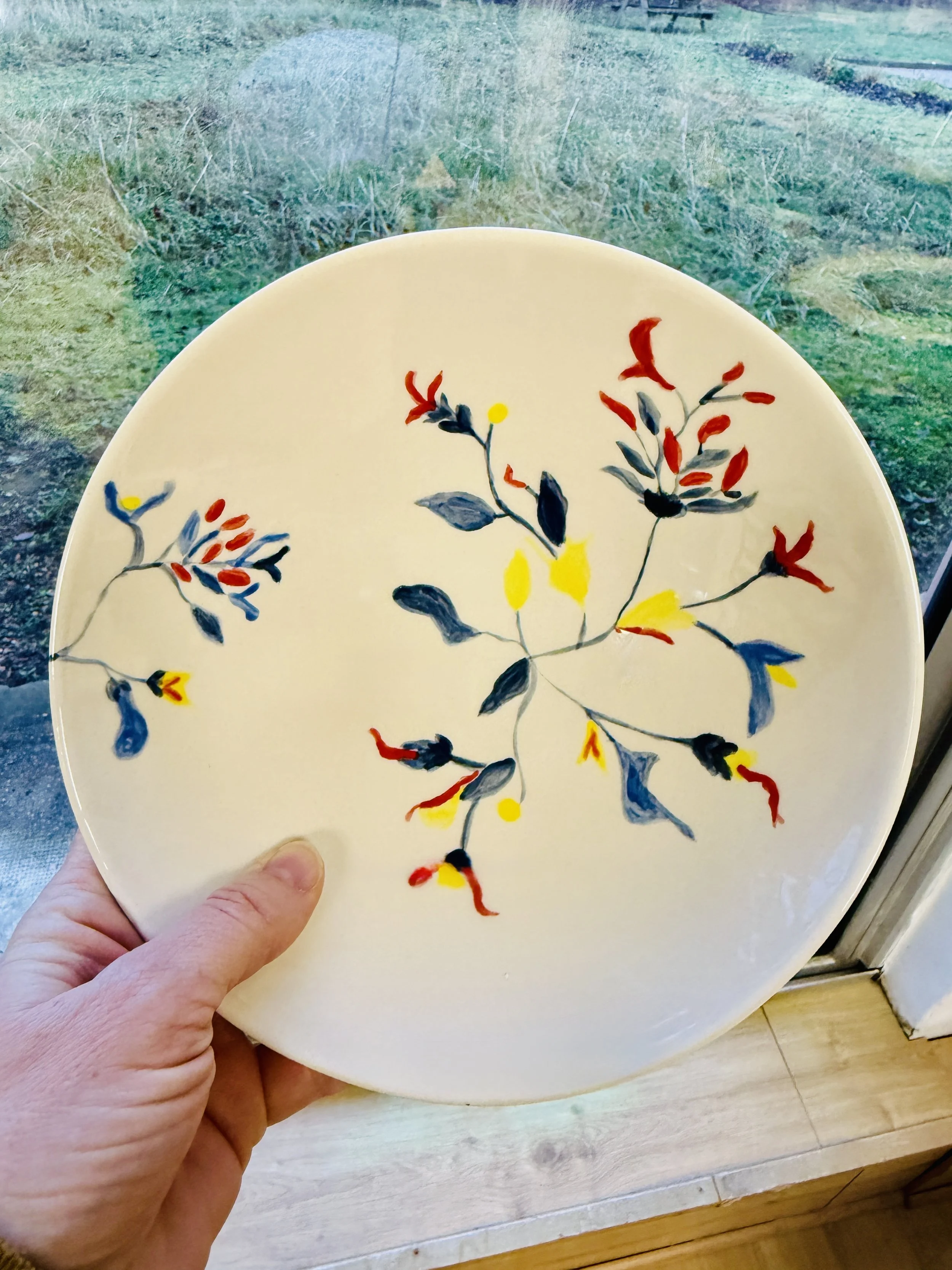 Assiette blanche avec un motif floral coloré en rouge, jaune, bleu et gris, tenue par une main, devant une fenêtre.