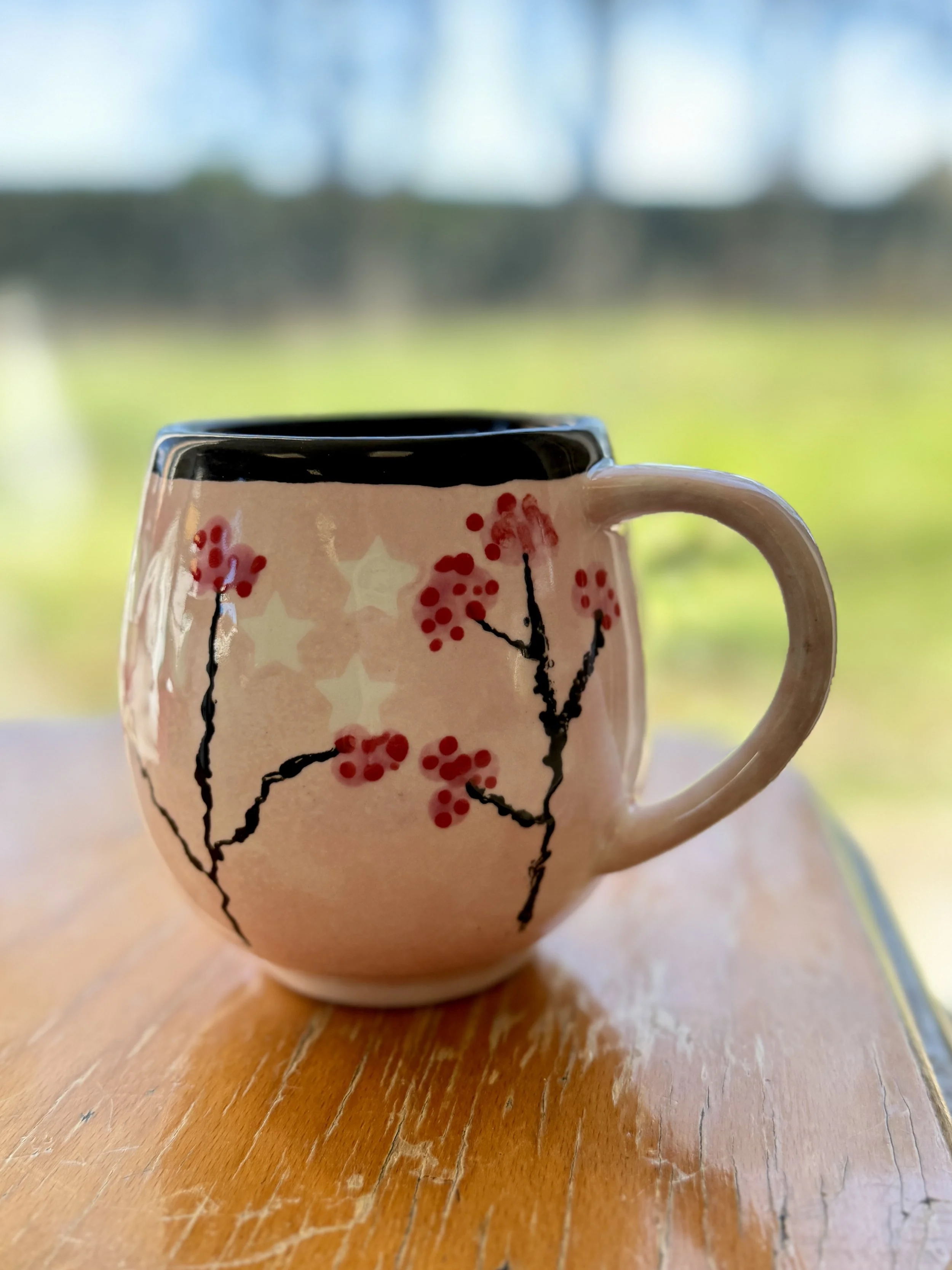 Une tasse en céramique décorée de branches de baies rouges, posée sur une table en bois avec un fond flou de paysage naturel.