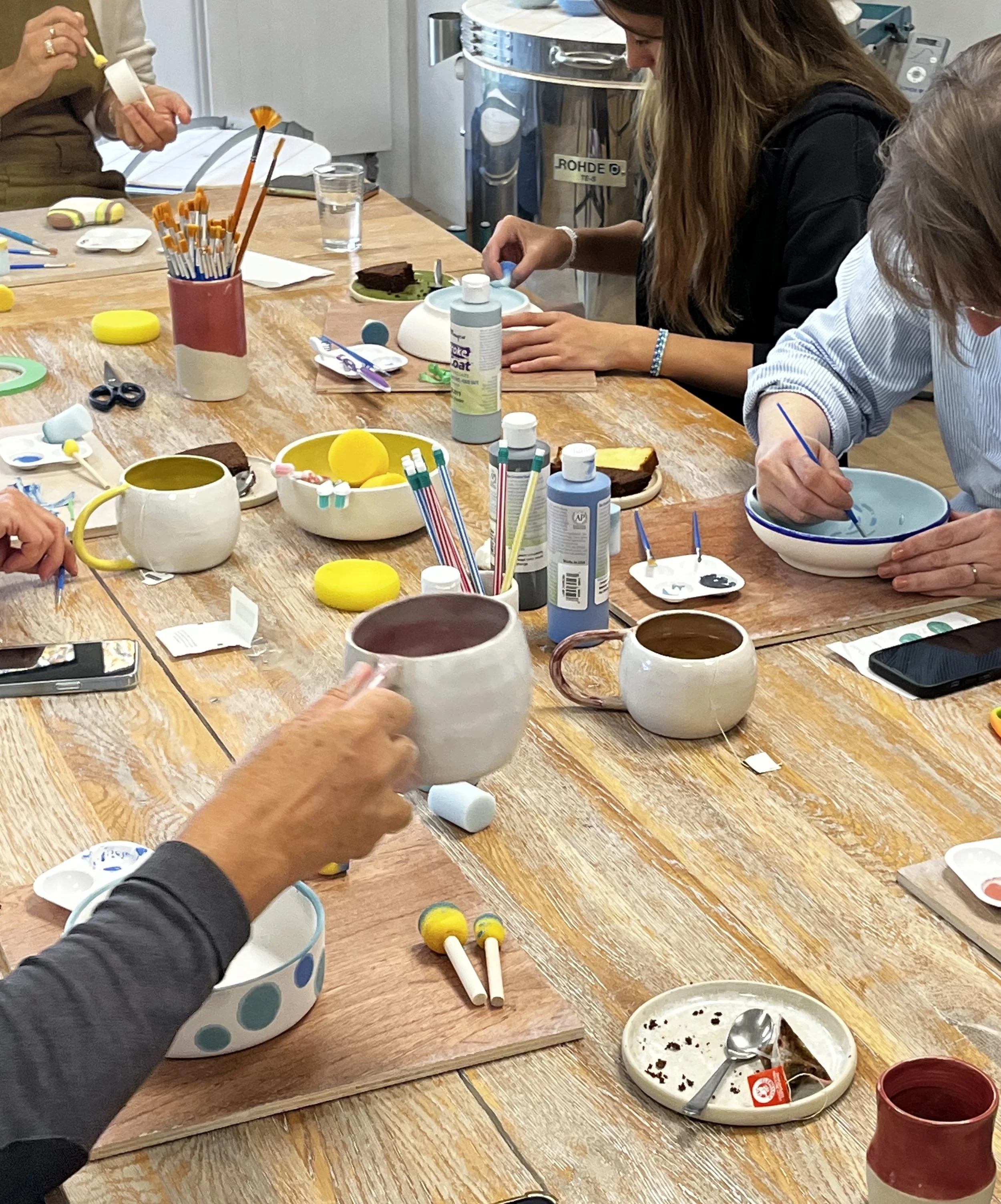 Groupe de personnes lors d'une activité de peinture sur céramique, utilisant des pinceaux et des peintures colorées sur des mugs et des bols en céramique sur une grande table en bois.