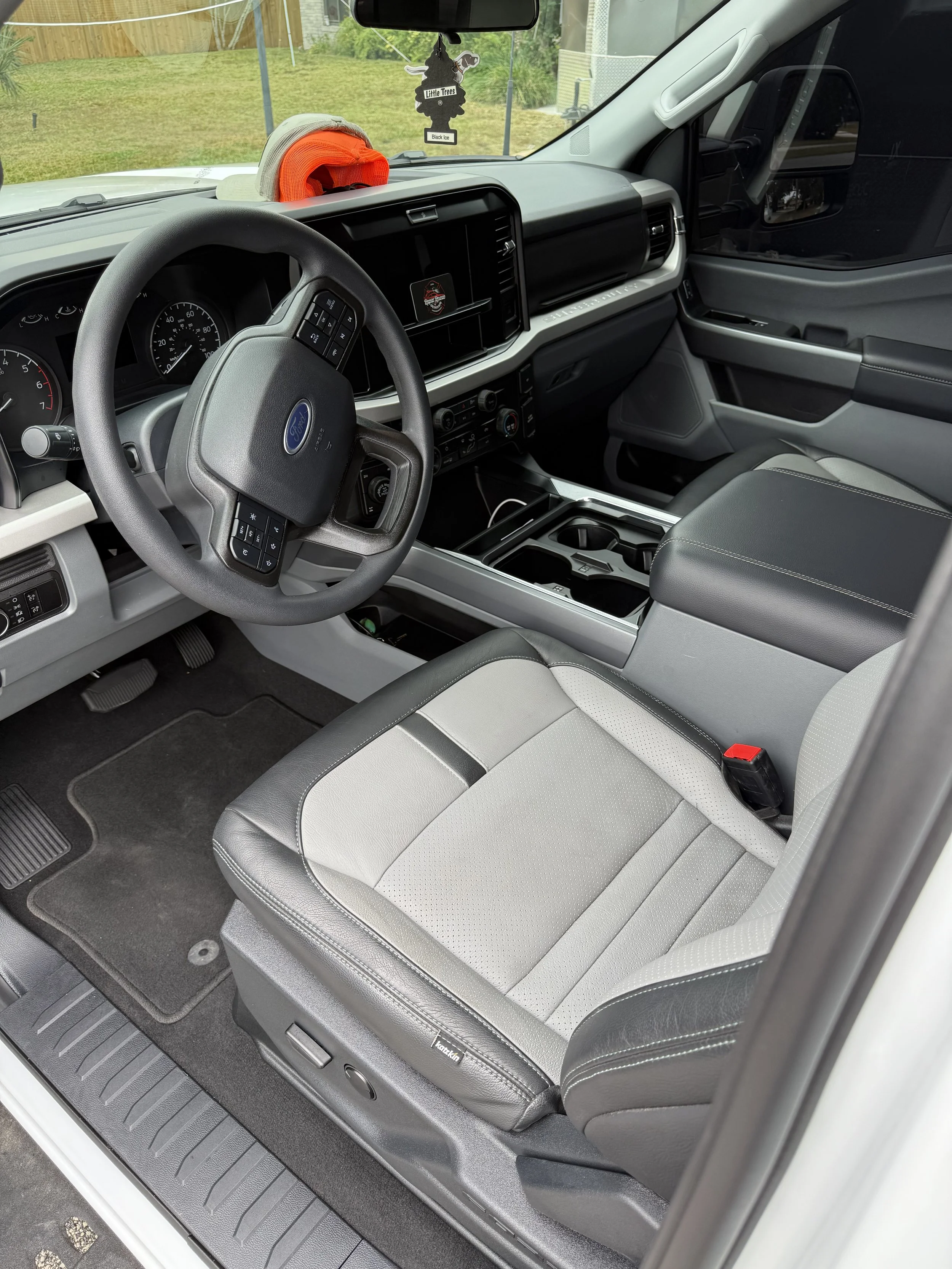 Interior of a Ford truck with a gray and black front seat, steering wheel, dashboard, and center console. There is a red cap, a hat, and an air freshener hanging from the rearview mirror.