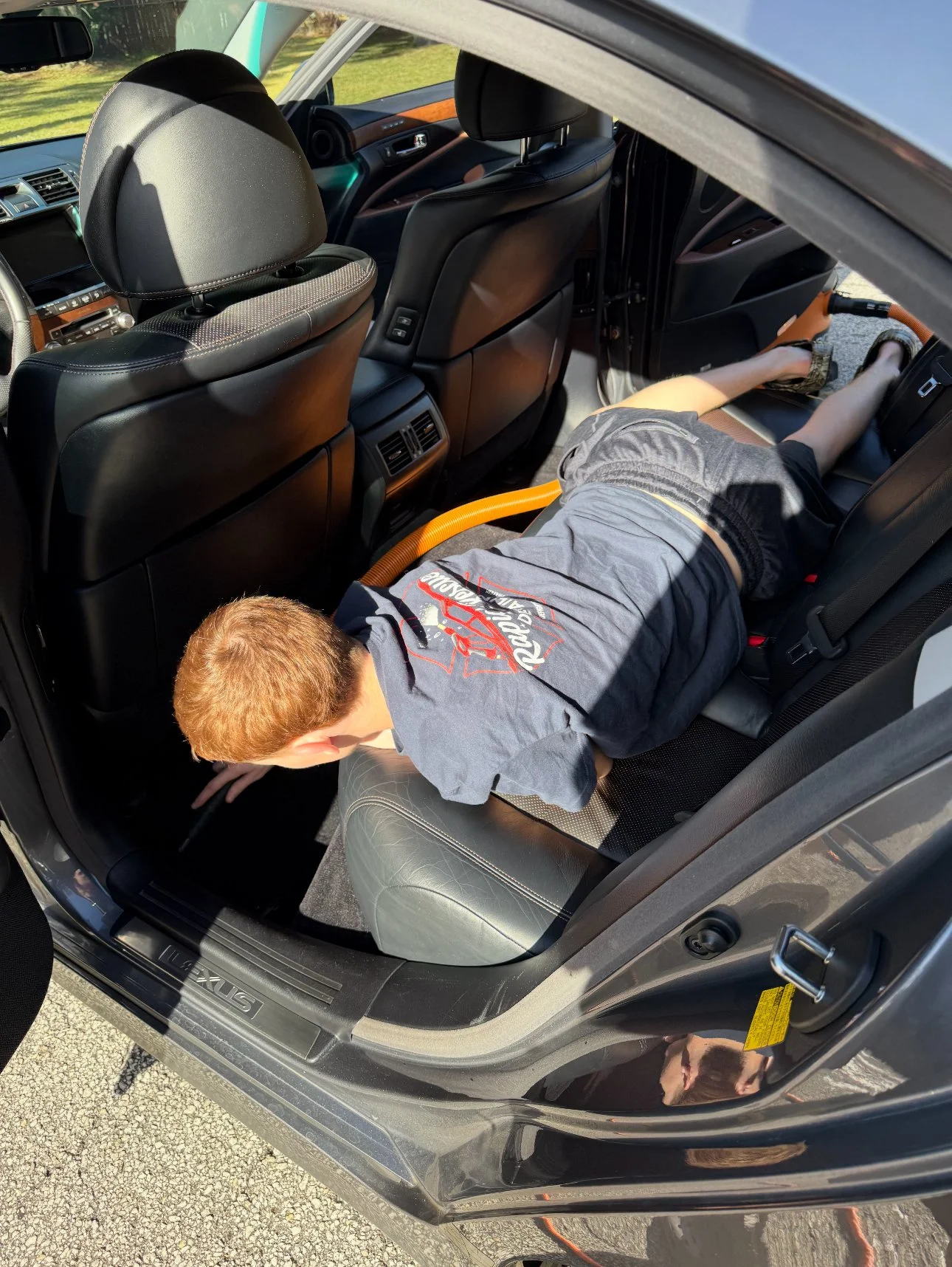 A young boy is leaning over in the back seat of a black car, reaching towards the floor. The interior of the car includes black leather seats and a small orange hose visible in the backseat area.