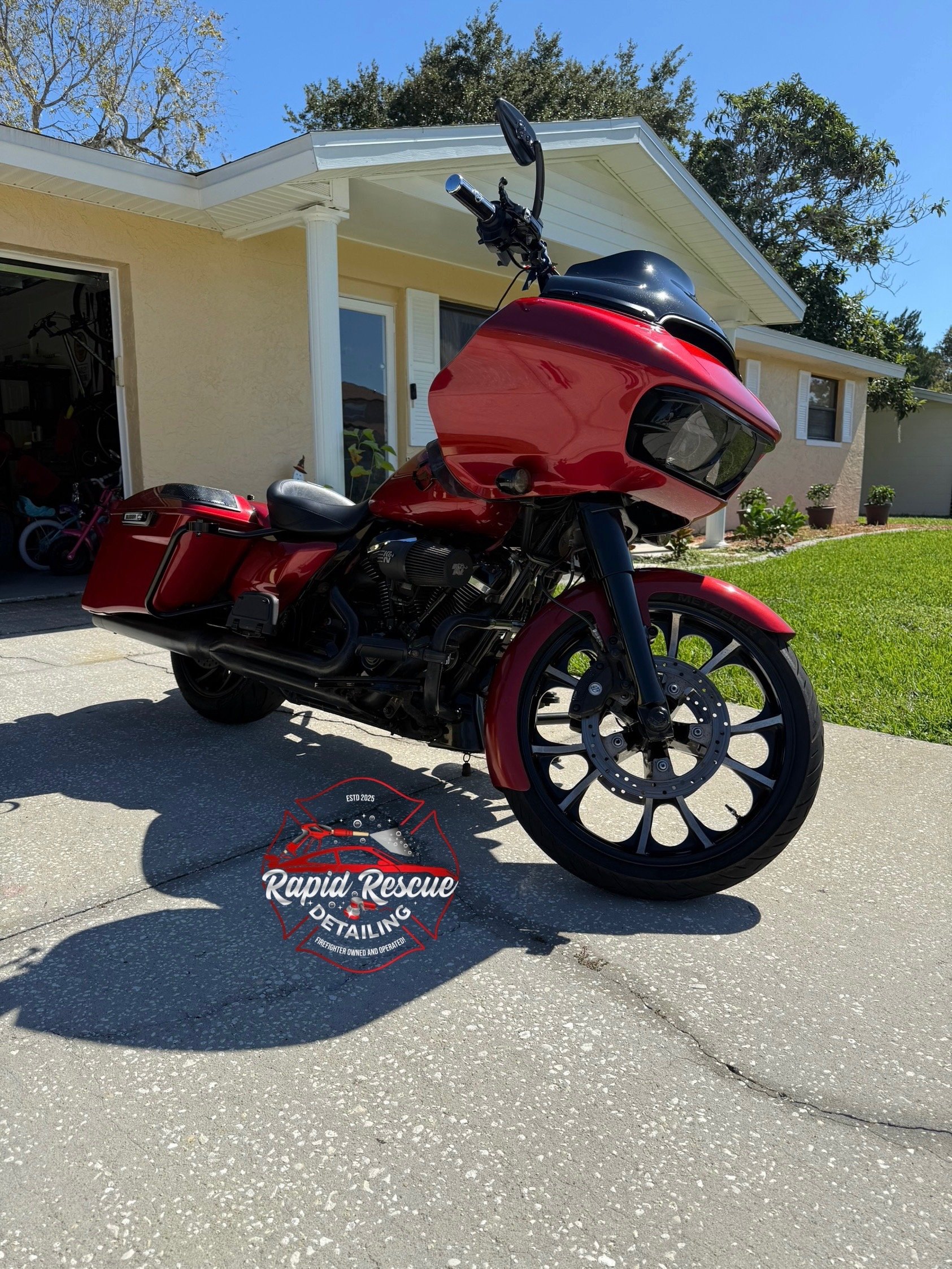 A red touring motorcycle parked on a driveway in front of a house, with a garage and a lawn in the background. There is a logo on the ground that reads 'Rapid Rescue Detailing'.