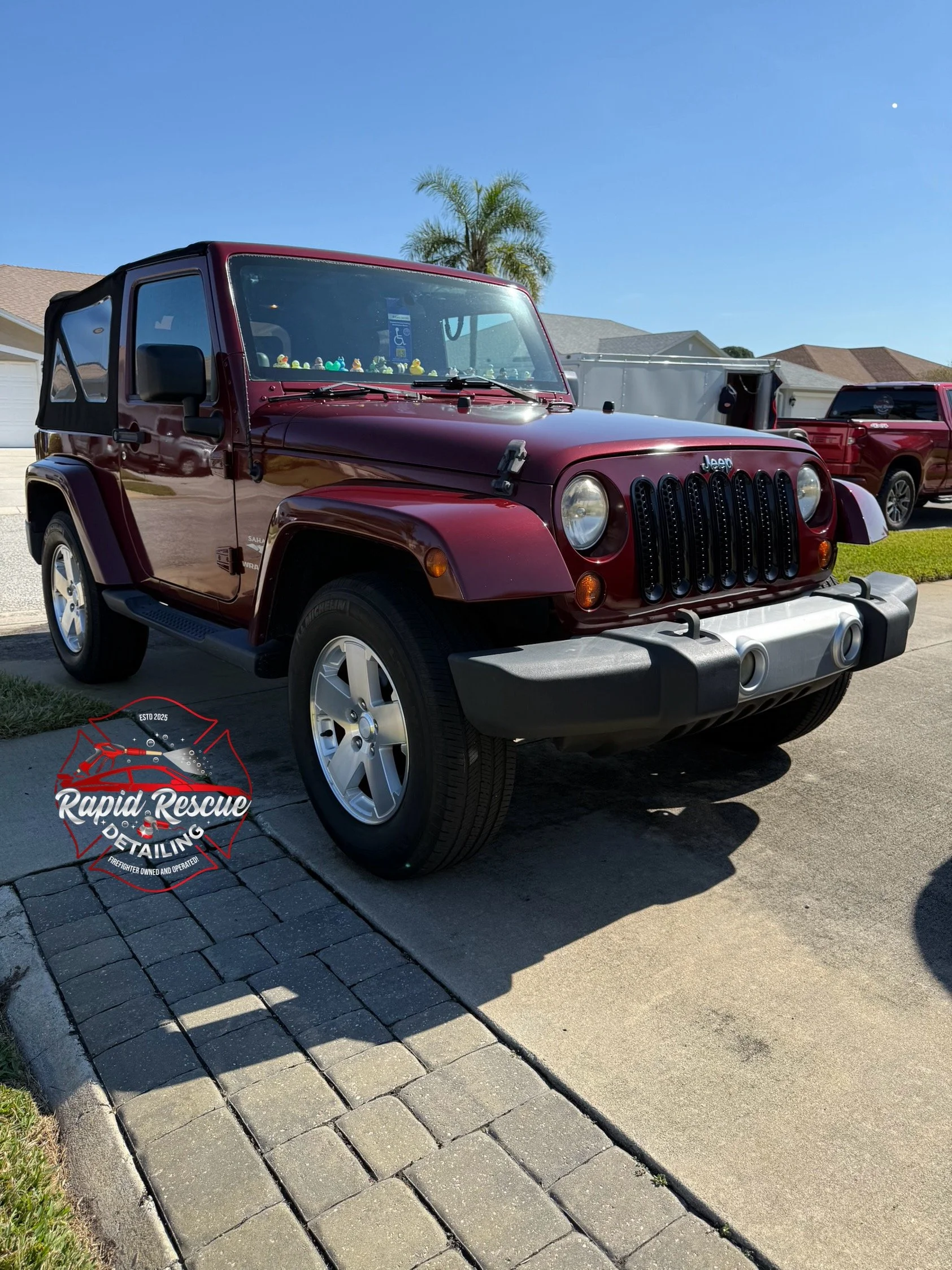 Maroon Jeep Wrangler parked on a driveway with a clear blue sky and a palm tree in the background.