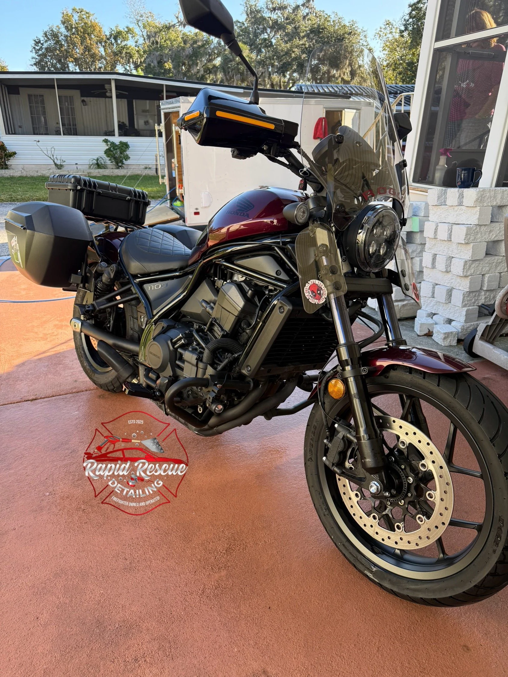 Black and red motorcycle parked outdoors on a reddish-tan concrete surface with a transparent windshield and saddlebags, nearby a house with white brick pillars and a porch, trees in the background.
