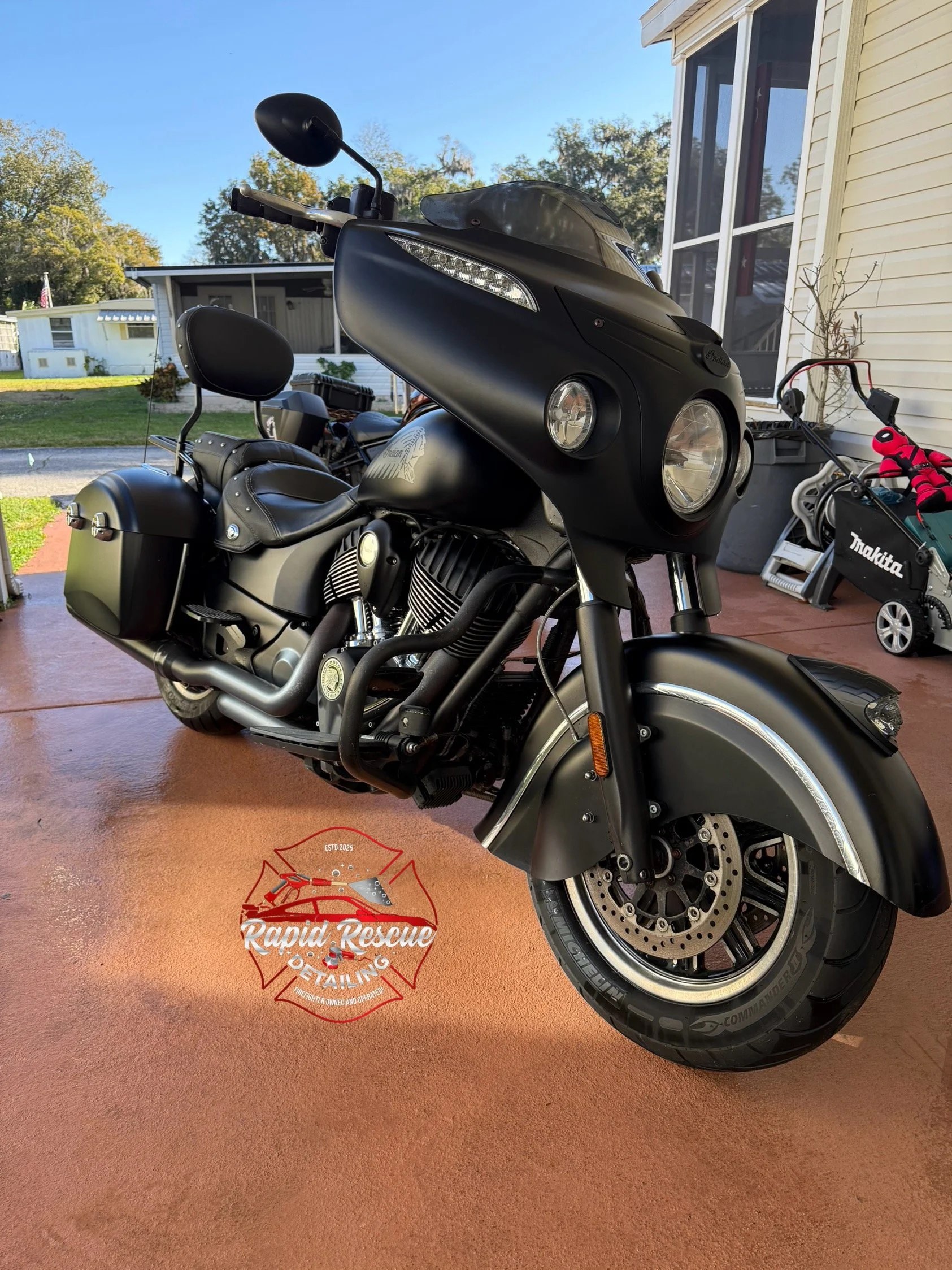 A black motorcycle parked on a red concrete driveway in a residential yard with trees and houses in the background. There is a logo on the ground that reads 'Rapid Rescue Detailing'.