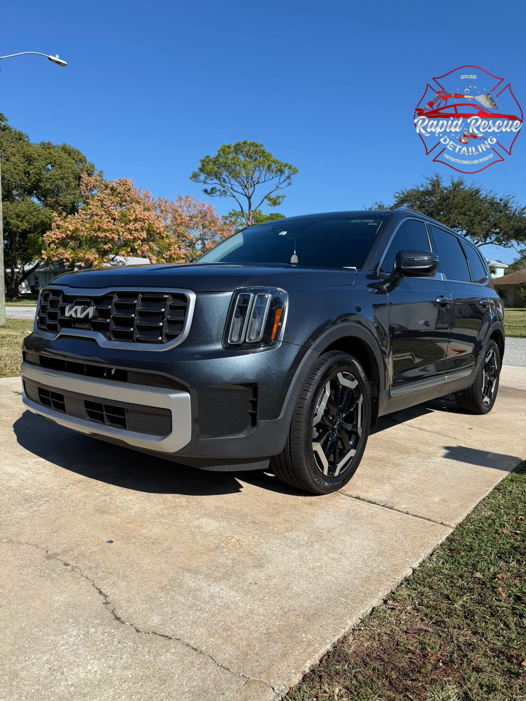 A dark gray Kia SUV parked on a concrete driveway with a suburban background, clear blue sky, trees, and a logo for Rapid Rescue Detailing in the upper right corner.