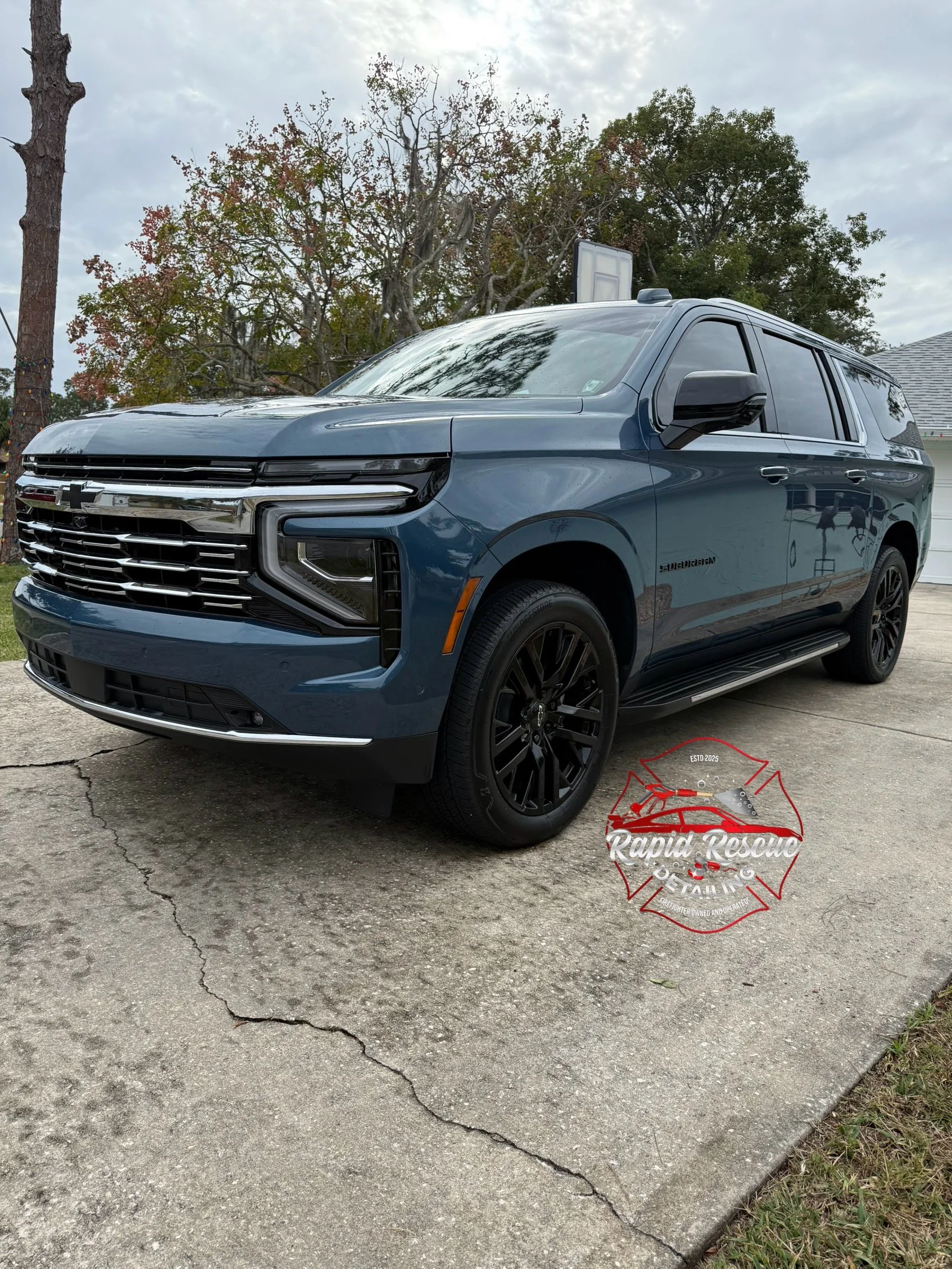 A blue Chevrolet Suburban parked on a cracked concrete driveway with trees in the background and a logo overlay for Rapid Rescue Detailing on the pavement.