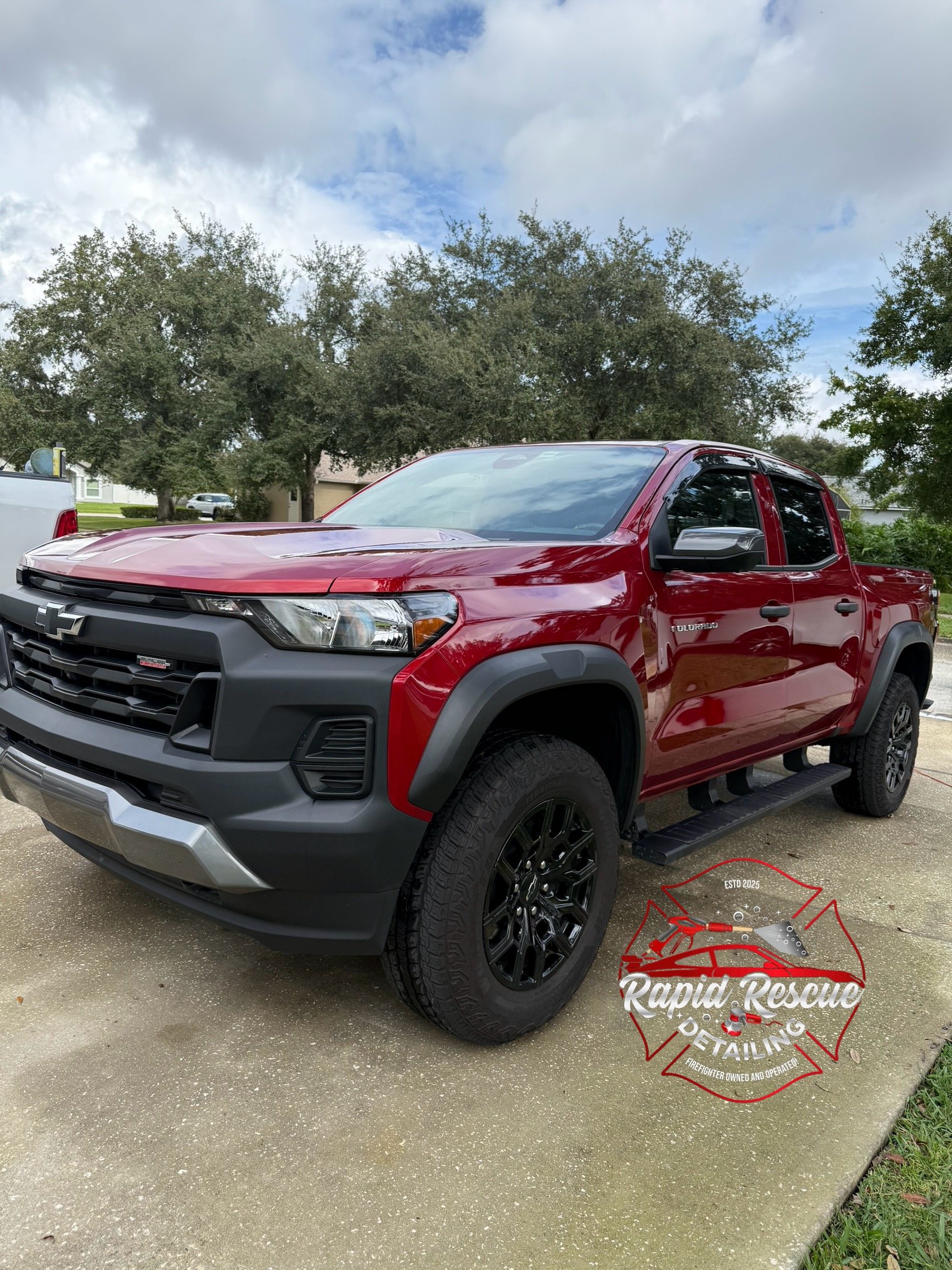 A red Chevrolet pickup truck parked on a driveway with trees and clouds in the background, and a logo for Rapid Rescue Detailing on the ground nearby.
