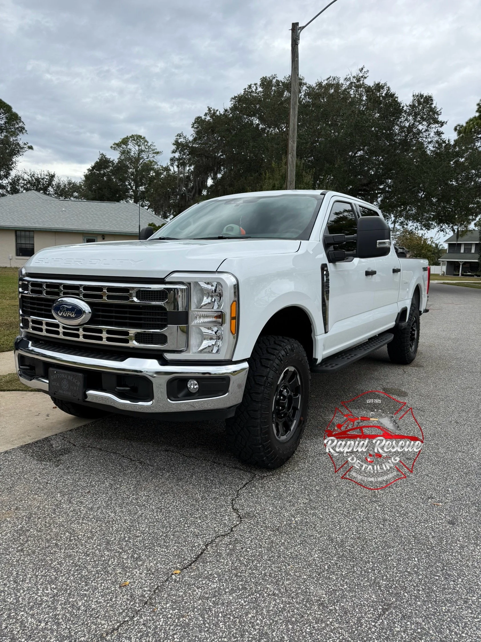 White Ford Super Duty pickup truck parked on a street with houses in the background and a cloudy sky. The truck has black wheels and a logo for Rapid Rescue Fire & Rescue.