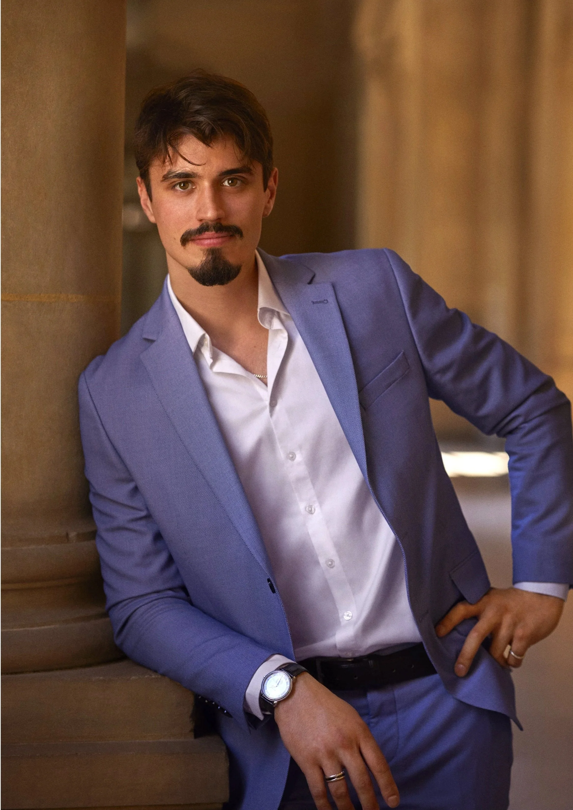 Un homme posa contre un mur en pierre, portant un costume bleu et une montre, dans un bâtiment architectural avec des colonnes, un palais de justice, un tribunal, une cour d'appel.