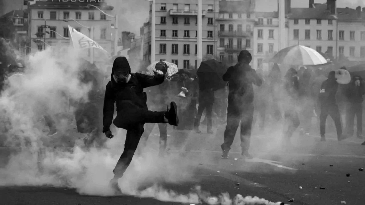 Manifestants et black bloc confrontant la police, avec de la fumée et des gaz lacrymogènes, dans une rue urbaine, certains portant des parapluies