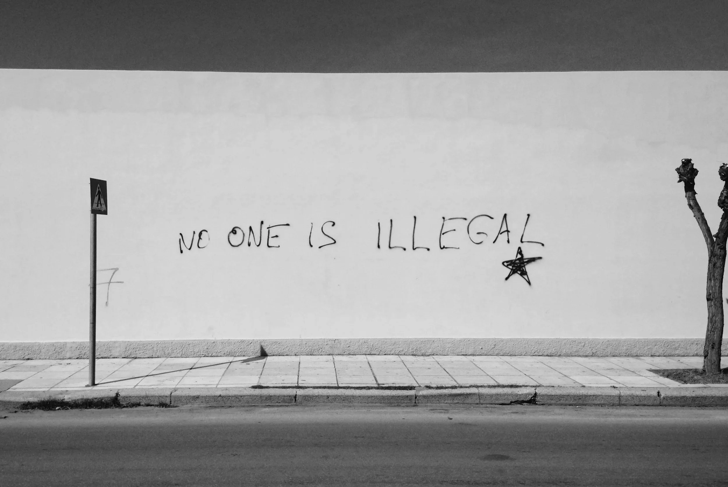 Mur blanc avec graffiti disant 'NO ONE IS ILLEGAL' et une étoile dessinée. Il y a un panneau piétonnier, un arbre sans feuilles, et une chaussée en bas.
Etrangers.