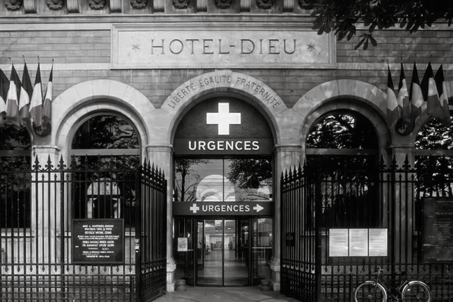 Entrée de l'urgence d'un hôpital, HOTEL-DIEU' à Paris avec des drapeaux français, un symbole de croix blanche, une grille en fer, et plusieurs affiches.