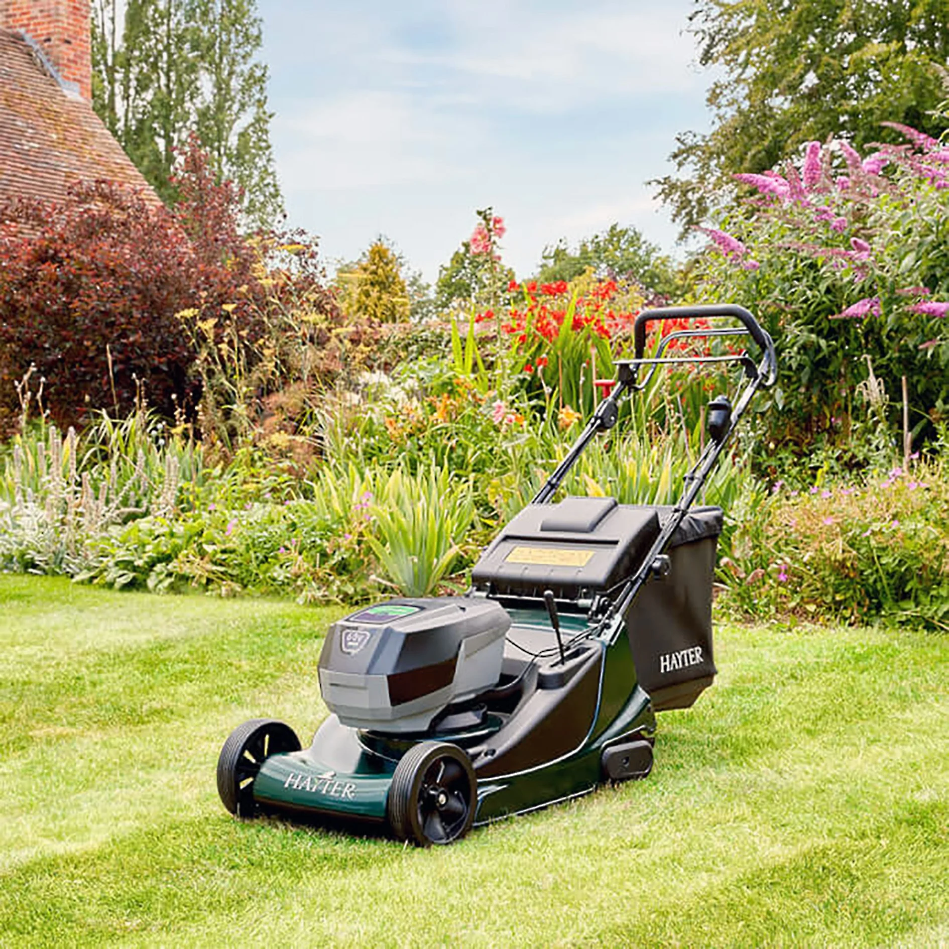 Electric lawn mower in a garden with colorful flowers and bushes, near a brick house wall.