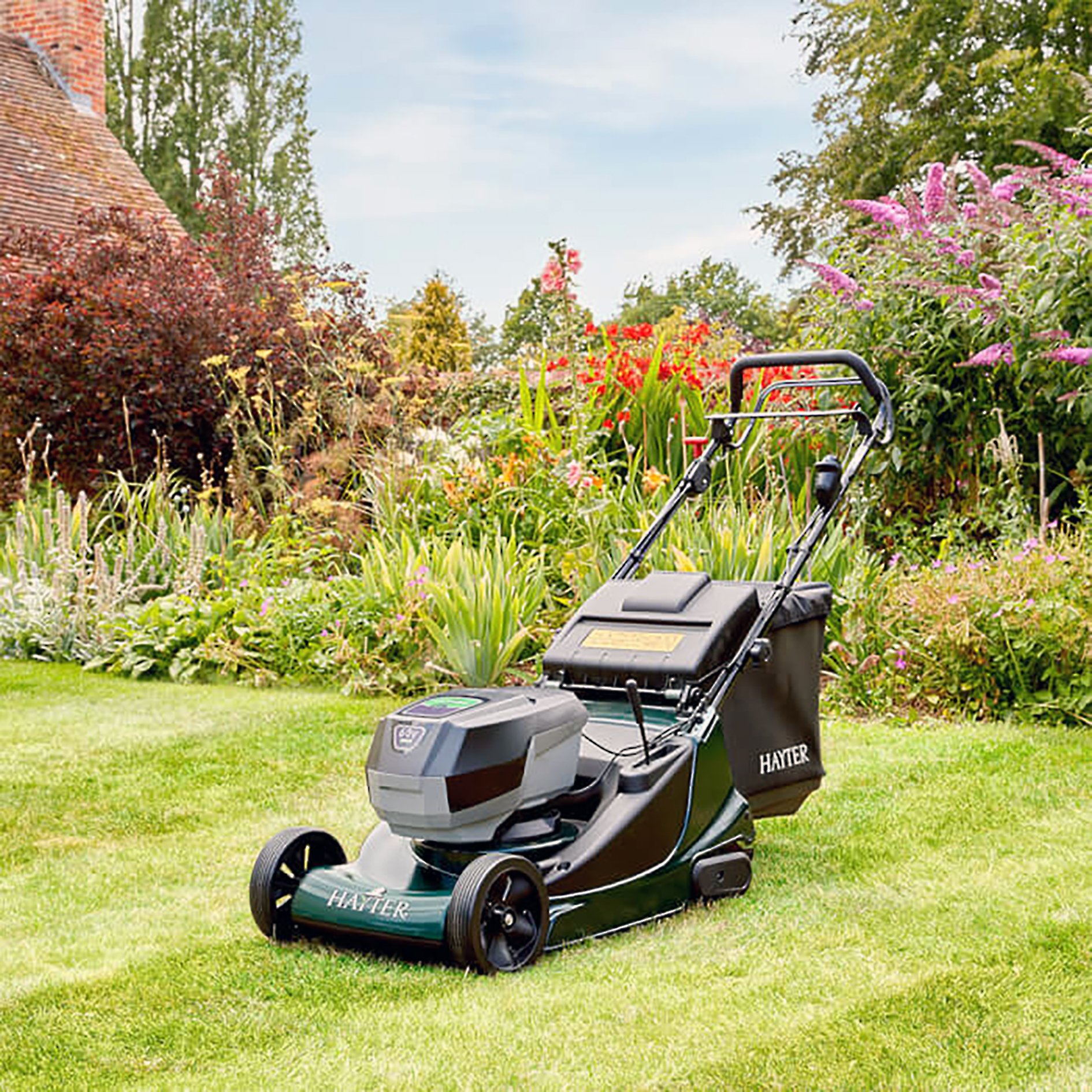 Electric lawnmower on a well-kept green lawn in a garden with colorful flowers and trees in the background.
