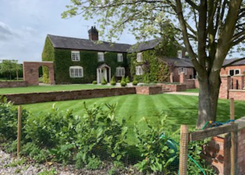 A large, ivy-covered brick house with multiple windows and a chimney, surrounded by a well-maintained lawn and garden, with a tree in the foreground and a brick wall in the background.