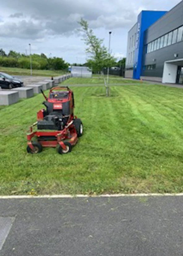 A red riding lawn mower on a well-maintained grassy area near a modern building with blue accents, a small leafless tree nearby, and parking spaces with cars in the background under a partly cloudy sky.