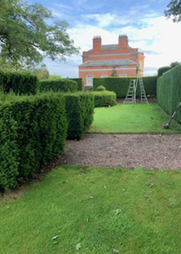 A neatly trimmed garden with a gravel pathway, green hedges, and a large brick house with chimneys in the background under a partly cloudy sky.