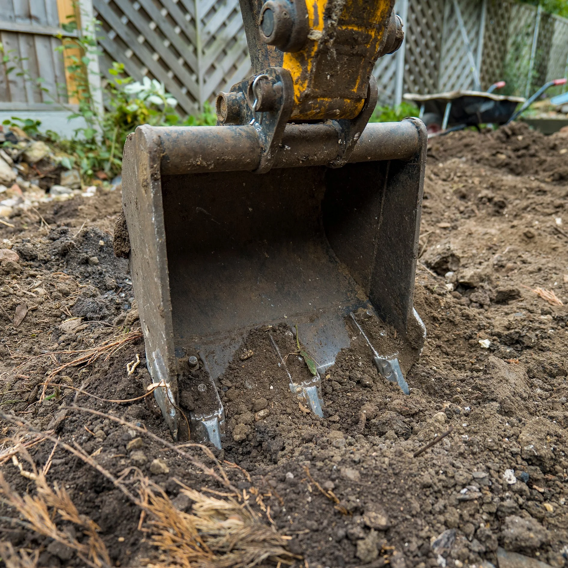 Close-up view of a small excavator bucket on dirt with a wooden fence and garden tools in the background.
