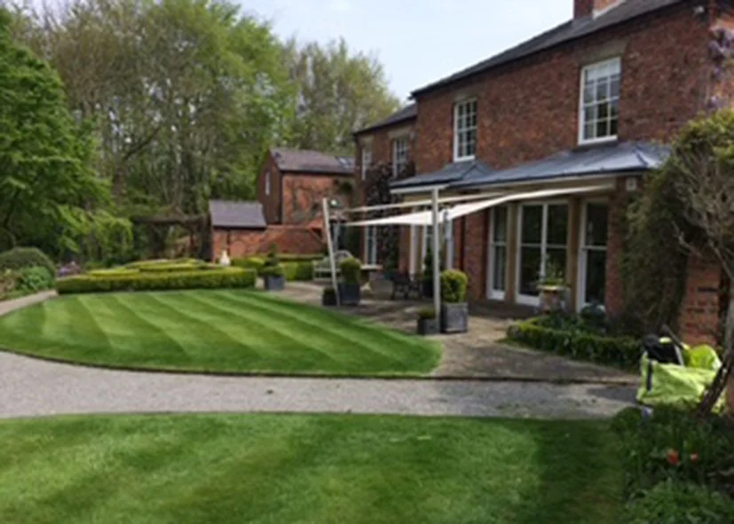 A brick house with a patio, surrounded by a well-manicured lawn, trees, and bushes.