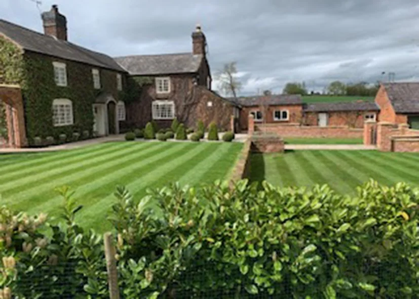 Large historic brick and stone mansion with neatly mowed striped lawn, shrubs, and a hedge in the foreground under a cloudy sky.