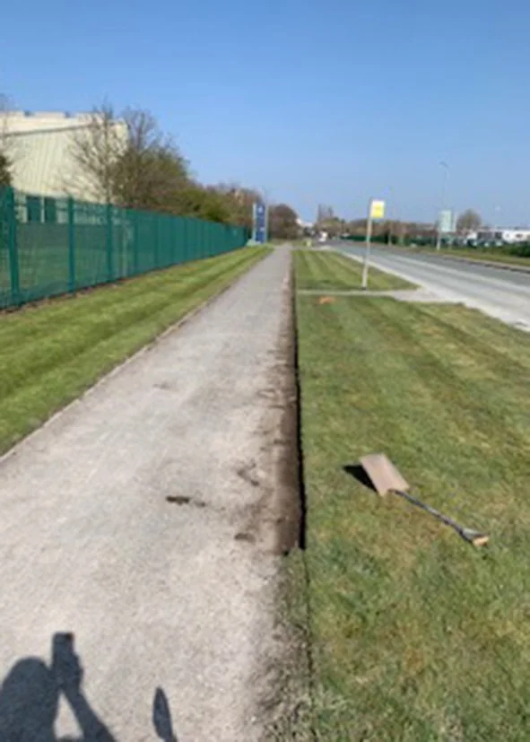 Sidewalk under construction on a sunny day with a shovel laying on freshly cut grass, a green fence on the left, and a busy road with cars on the right.