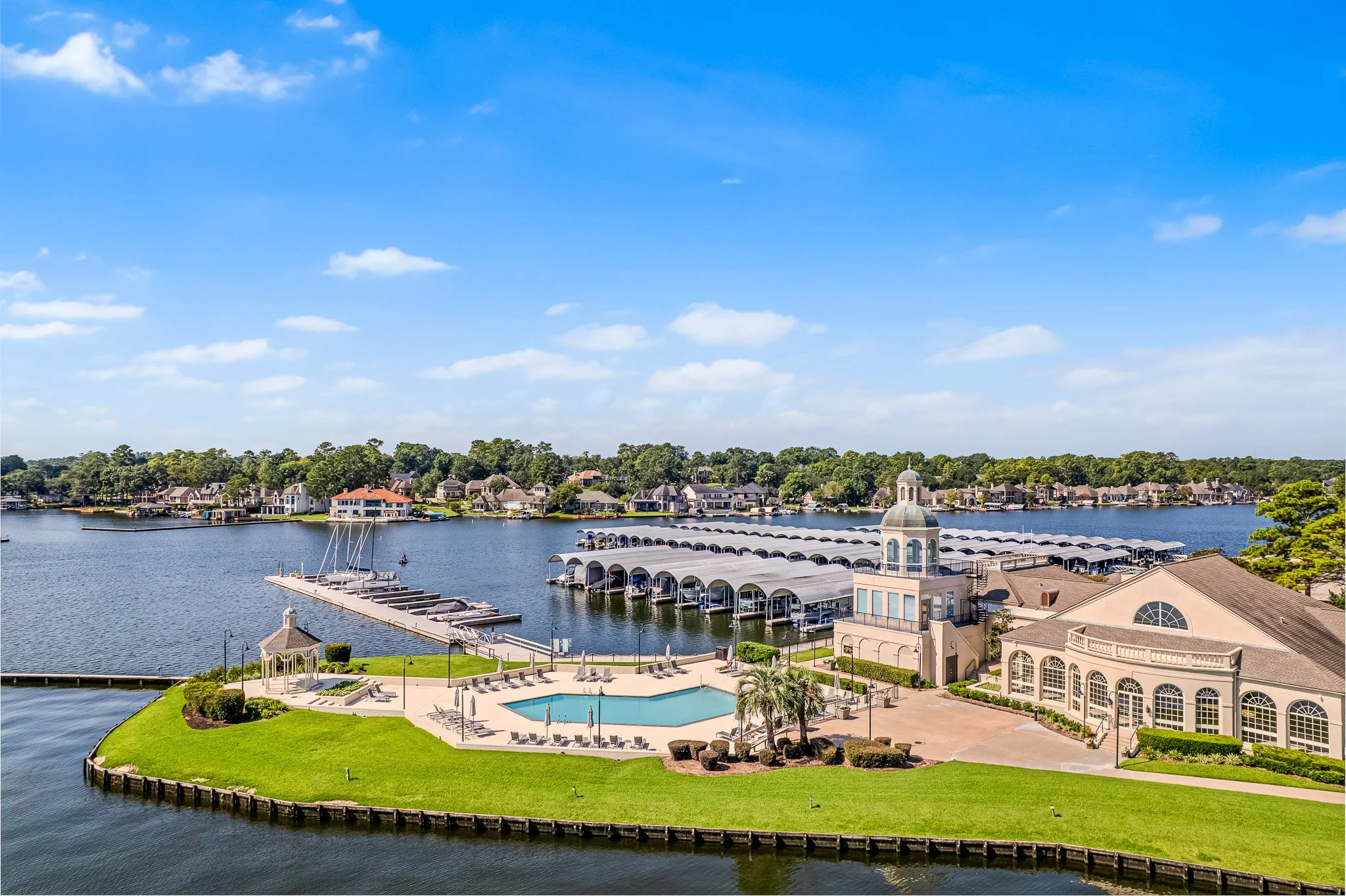 A lakeside residential community with a clubhouse, swimming pool, and marina. The scene includes houses along the water, a dock with boats, and a boat storage area with covered parking. The sky is blue with some clouds.