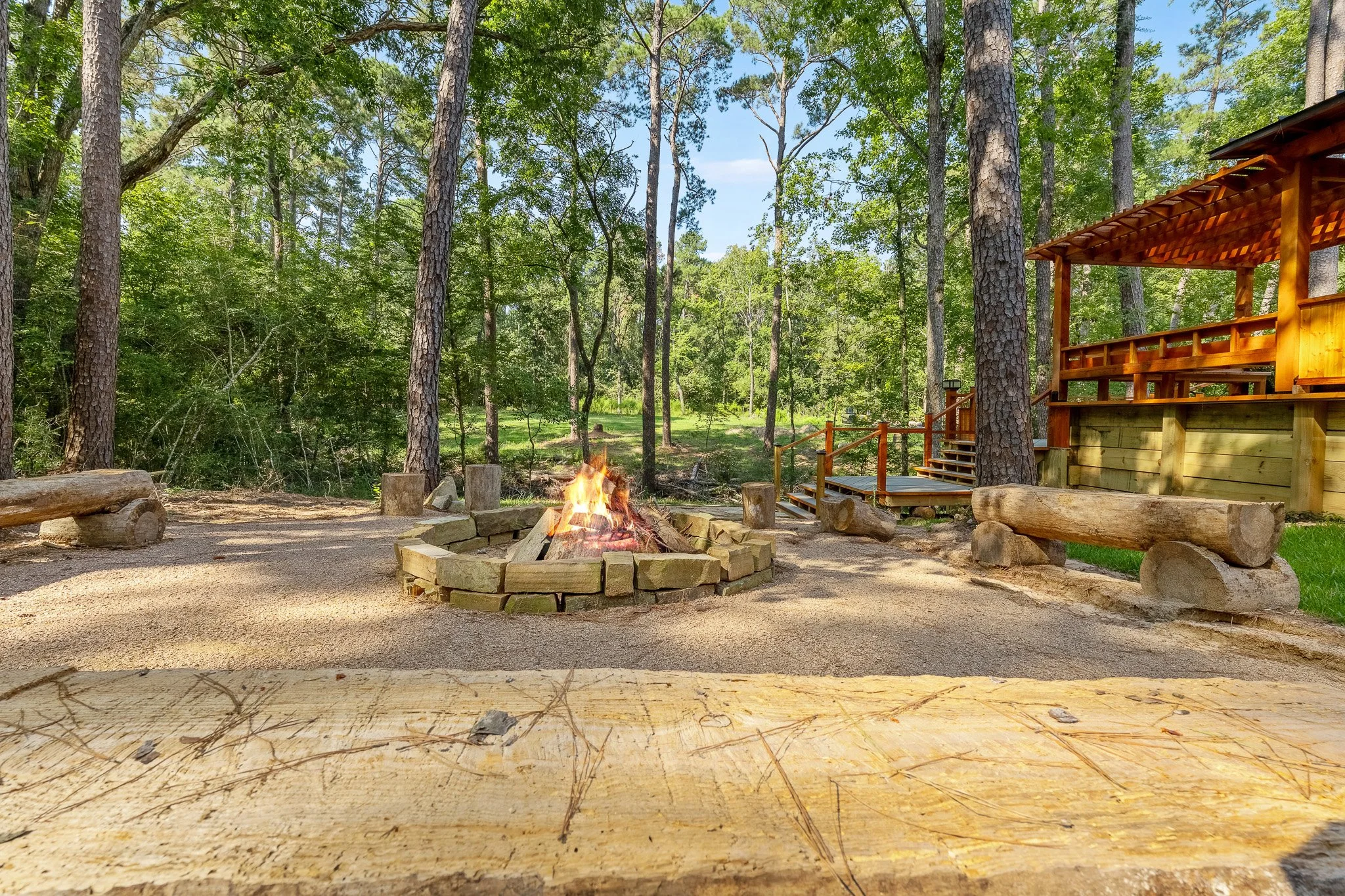 Wooden benches and a fire pit in a forested outdoor area with trees and a wooden deck.