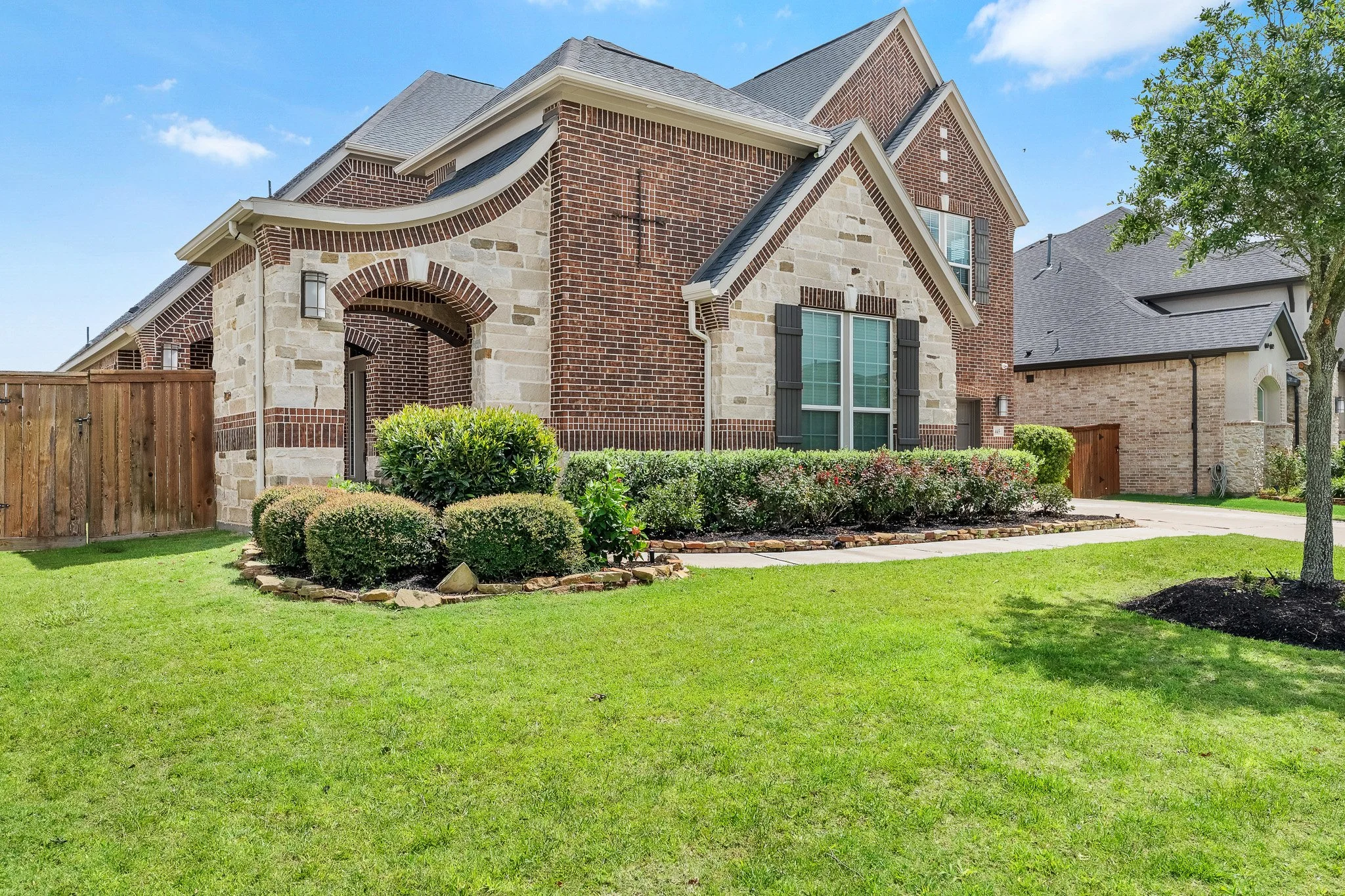 Front yard of a brick and stone house with green lawn, landscaped bushes, and a sidewalk, under a partly cloudy blue sky.