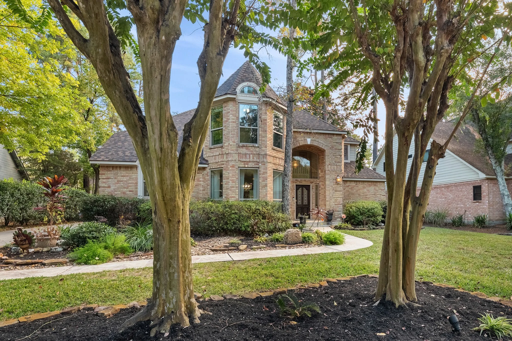 A brick house with a front entrance framed by trees and a well-maintained yard, including shrubs, plants, and a curved walkway.