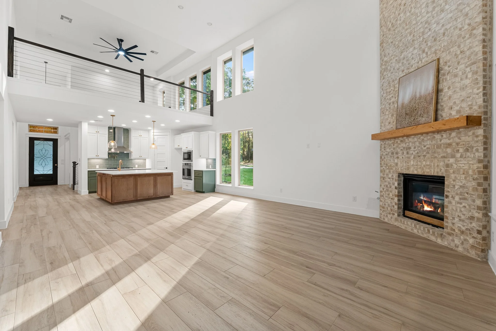 Open-concept living room and kitchen area in a modern home with high ceilings, large windows, a fireplace, wood flooring, and minimal decor.