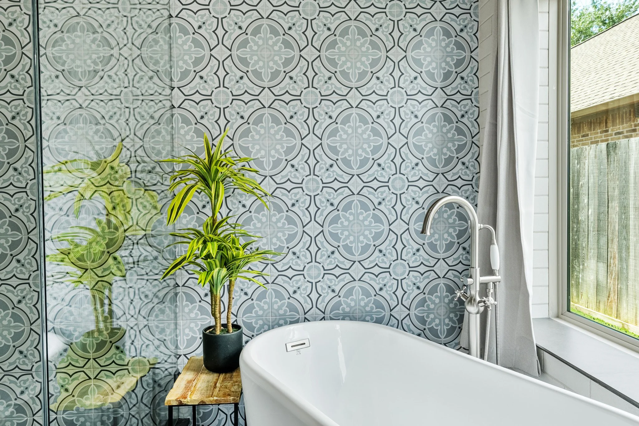 Bathtub next to a window with light gray curtains, a plant on a wooden table, and a wall with gray and white patterned tile.