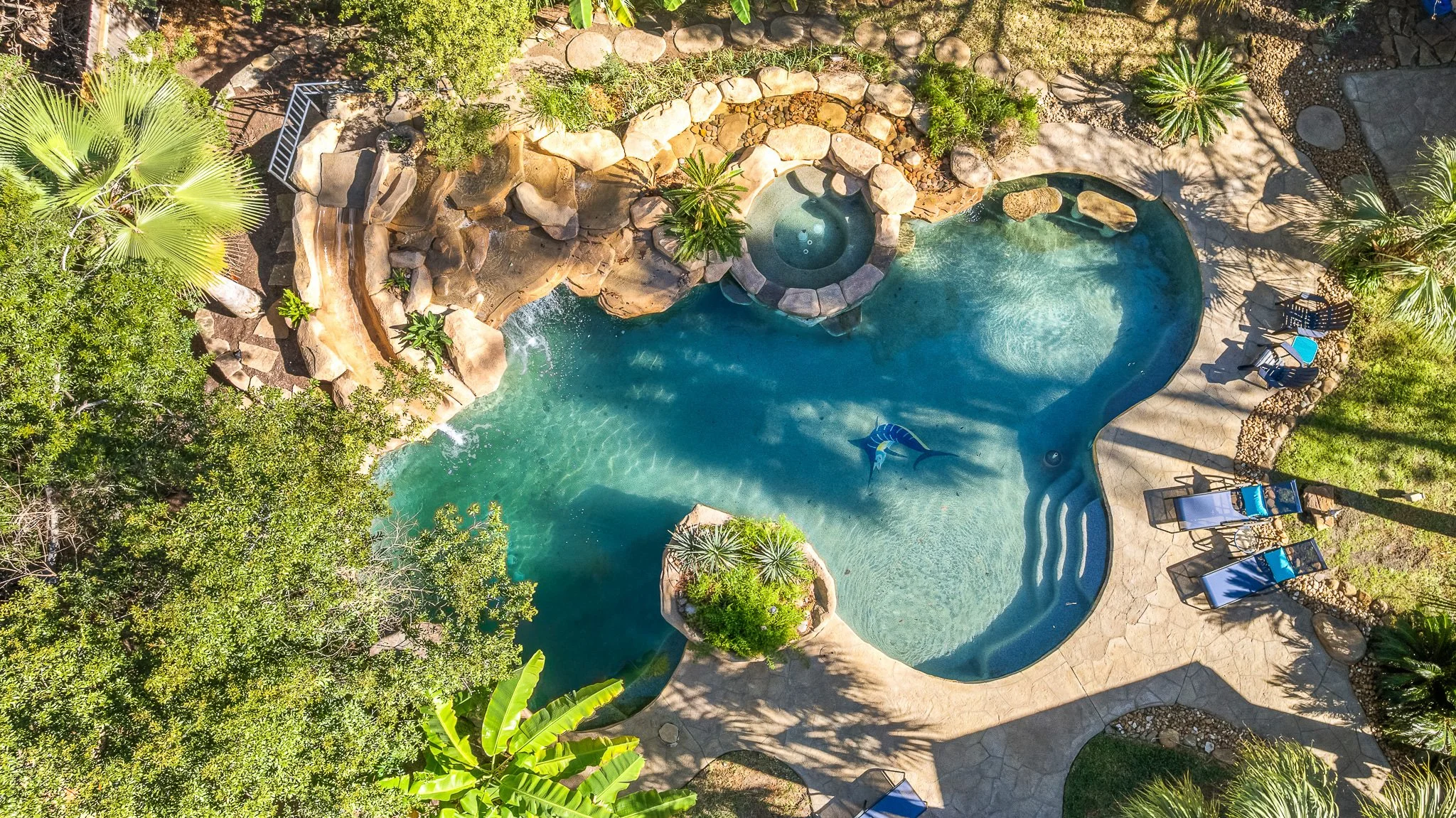 An aerial view of a backyard swimming pool surrounded by tropical plants, trees, and stone landscaping, with lounge chairs, umbrellas, and a hot tub nearby.