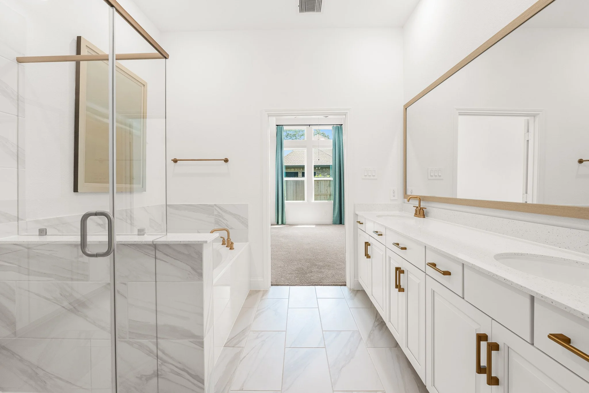 Bathroom with white cabinets, gold hardware, large mirror, and a window with teal curtains, leading to a carpeted room.
