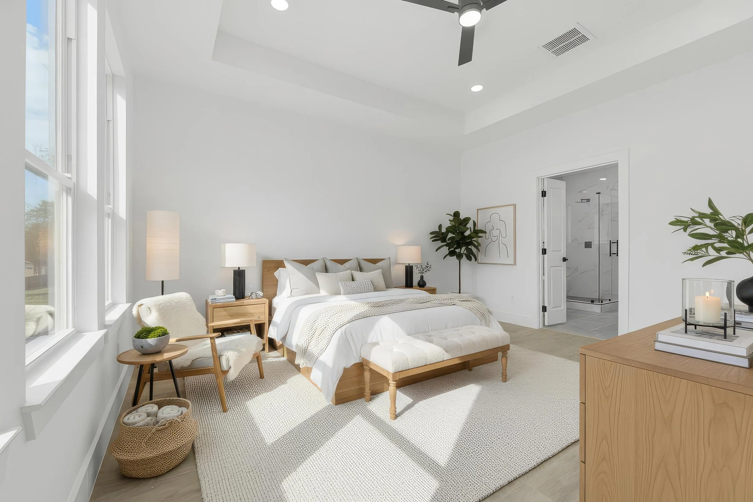 A bright, modern bedroom with white walls, large windows, a wooden bed with white bedding, a bench at the foot of the bed, and a beige rug. There are nightstands with lamps and decor items, and an open door leads to a bathroom with a glass shower.