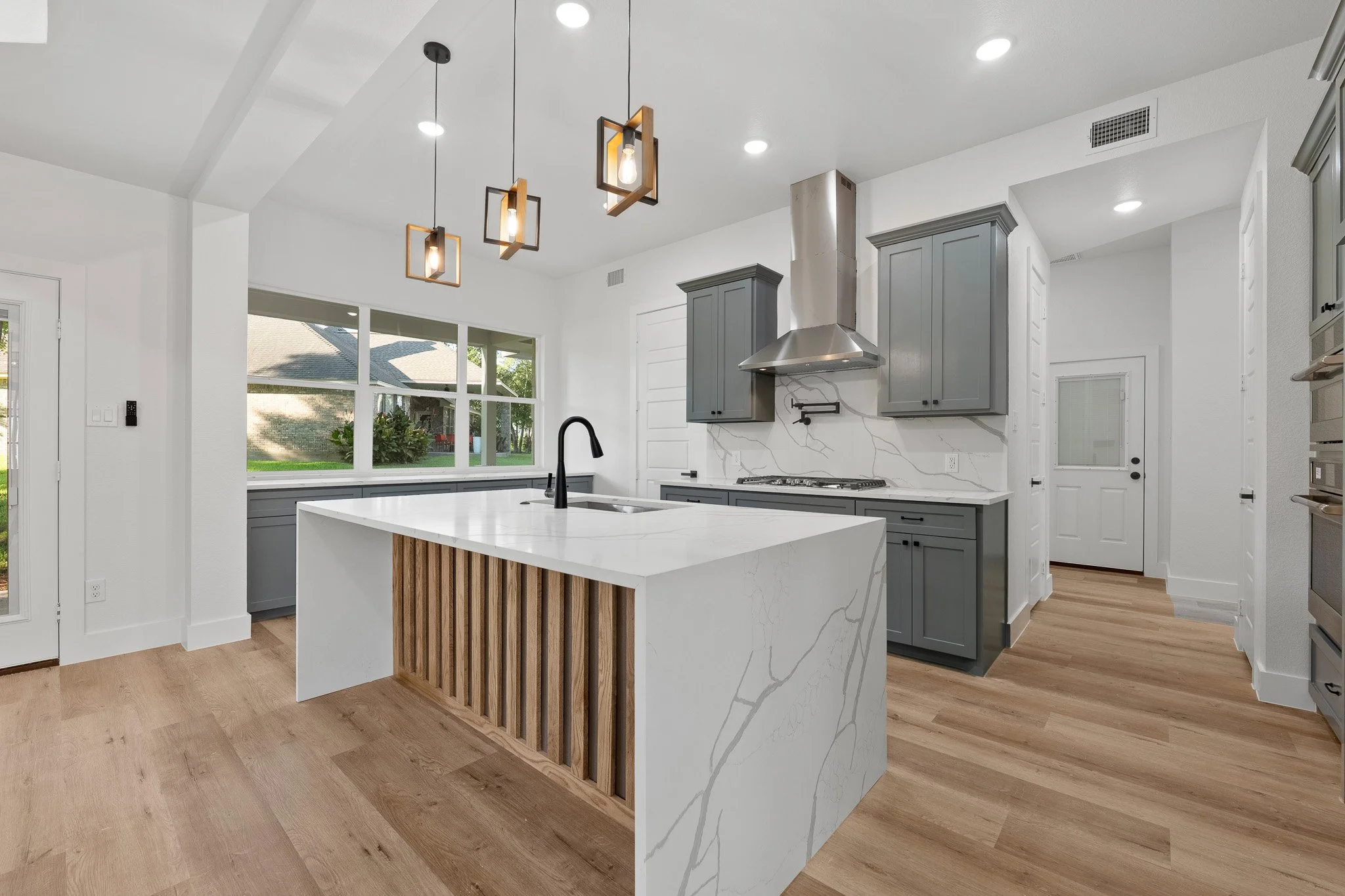 Modern kitchen with white and gray cabinetry, a marble island with wooden accents, large windows, pendant lights, and a hardwood floor.