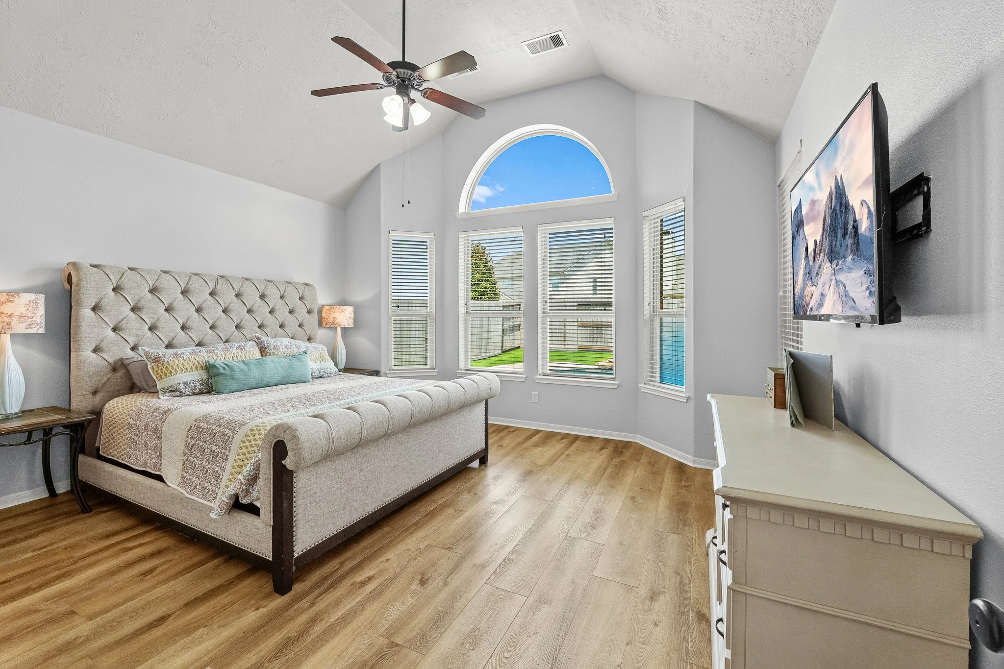 Bright bedroom with a large tufted beige bed, two bedside lamps, a white dresser, and a wall-mounted TV. Large arched window and several smaller windows let in natural light, showing a backyard with a fence and a tree. Wood floor and ceiling fan with