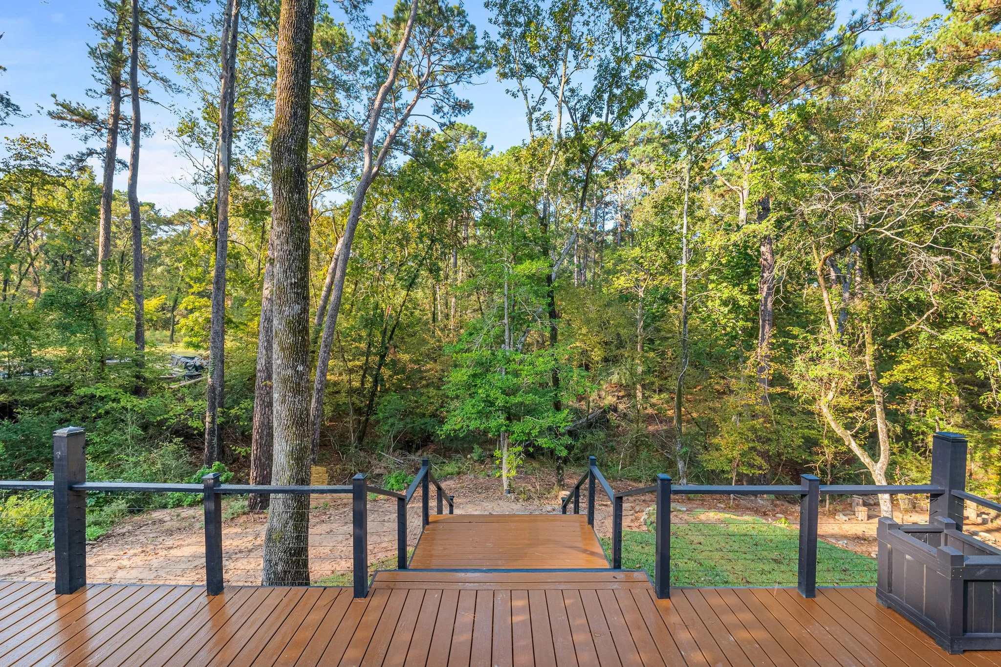 A wooden deck with black railings overlooking a lush green forest with tall trees and sunlight filtering through leaves.