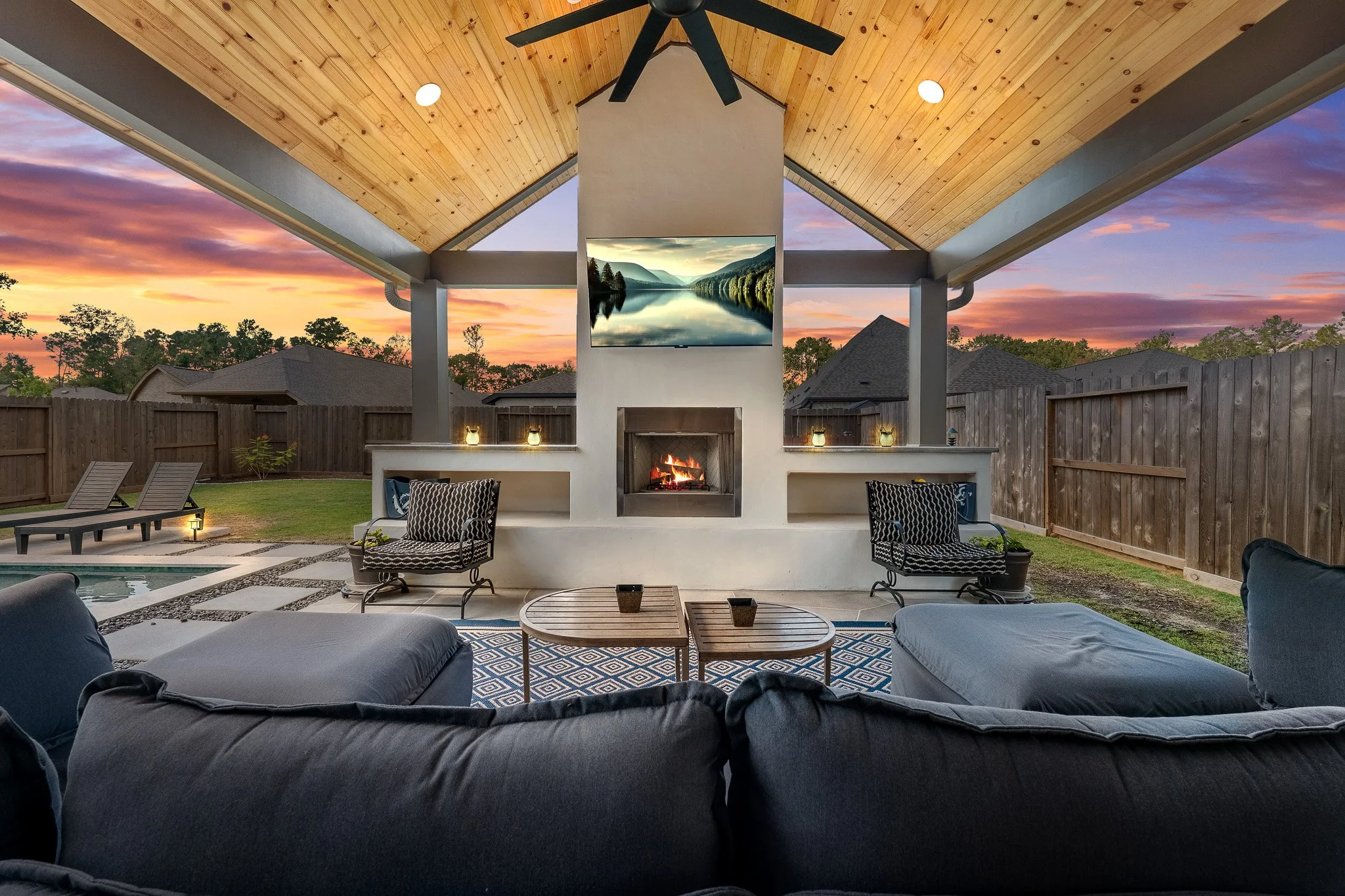 Outdoor living space featuring a covered patio with a wooden ceiling, a central fireplace, a wall-mounted TV, and outdoor furniture, with a backyard, lounge chairs, and a sunset sky in the background.