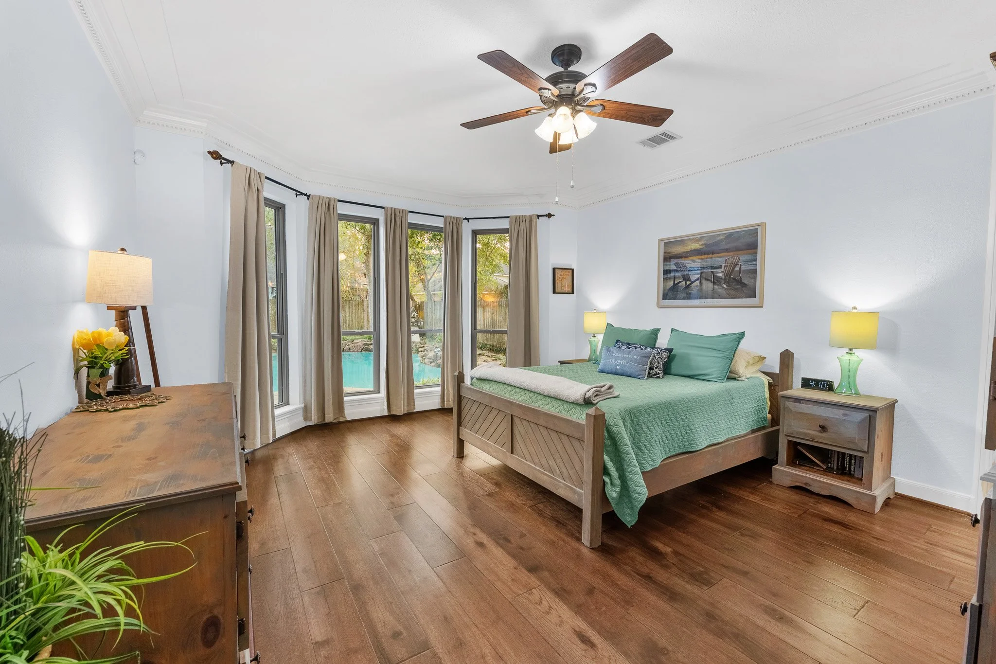A bedroom with a large bed, green bedding, and multiple pillows, next to a nightstand with a lamp, and a wall art above the bed. There are windows with beige curtains, and a view of a backyard with a swimming pool.