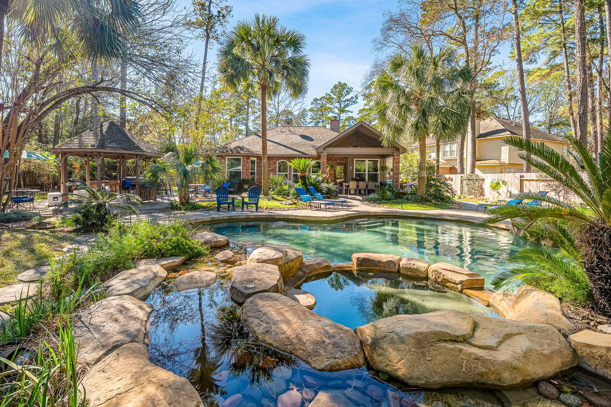 Backyard with a swimming pool surrounded by palm trees, lounge chairs, a gazebo, and a house in the background.