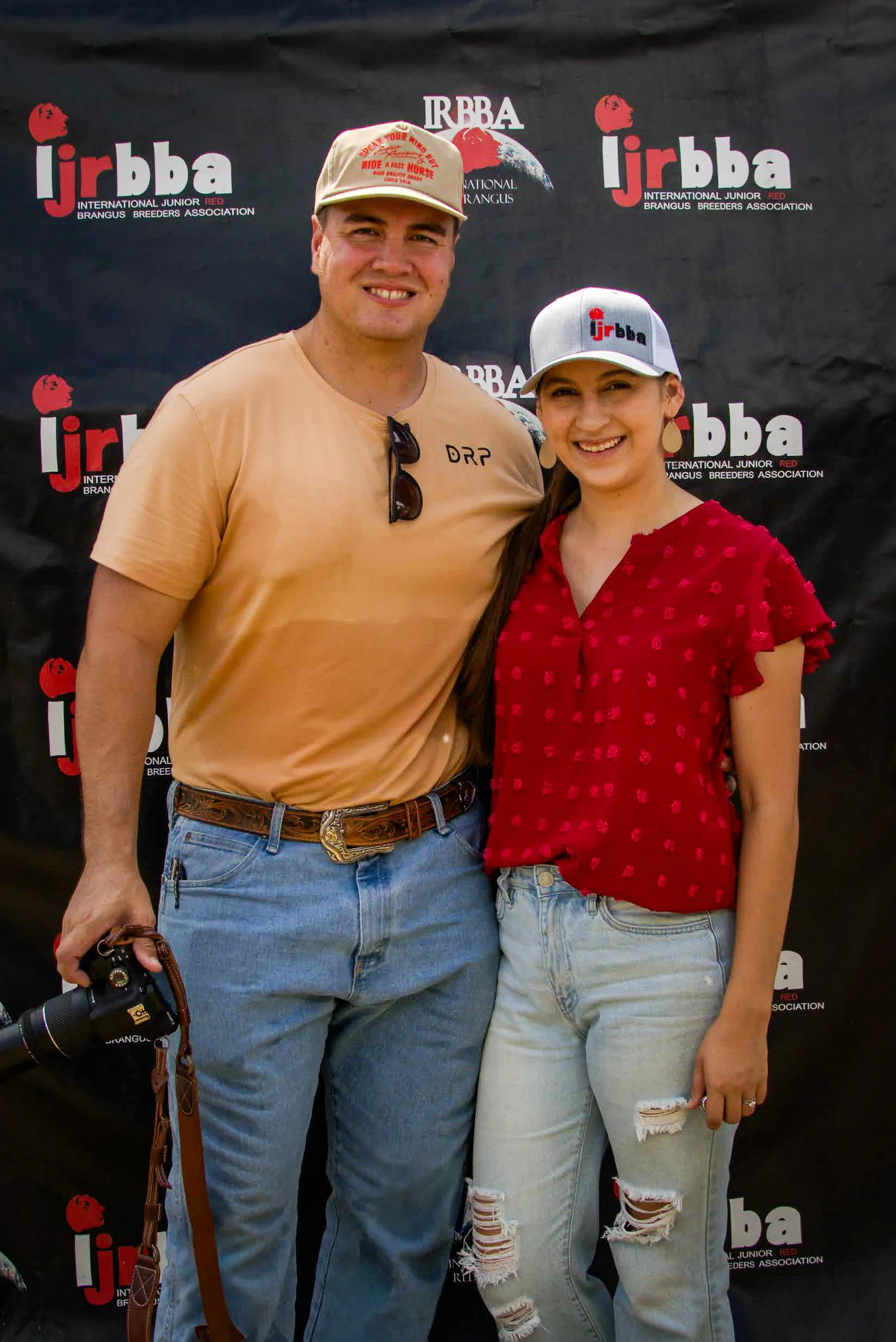 Two people standing together in front of a black backdrop with the logo of the International Junior Red Brangus Breeders Association. The man is wearing a beige hat, tan T-shirt, and jeans, holding a camera. The woman is wearing a white cap, red blouse, and ripped jeans.