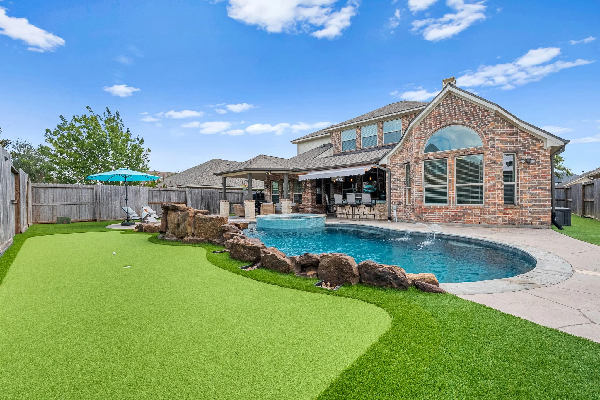 Backyard with a swimming pool, artificial grass, and a patio with a house in the background.