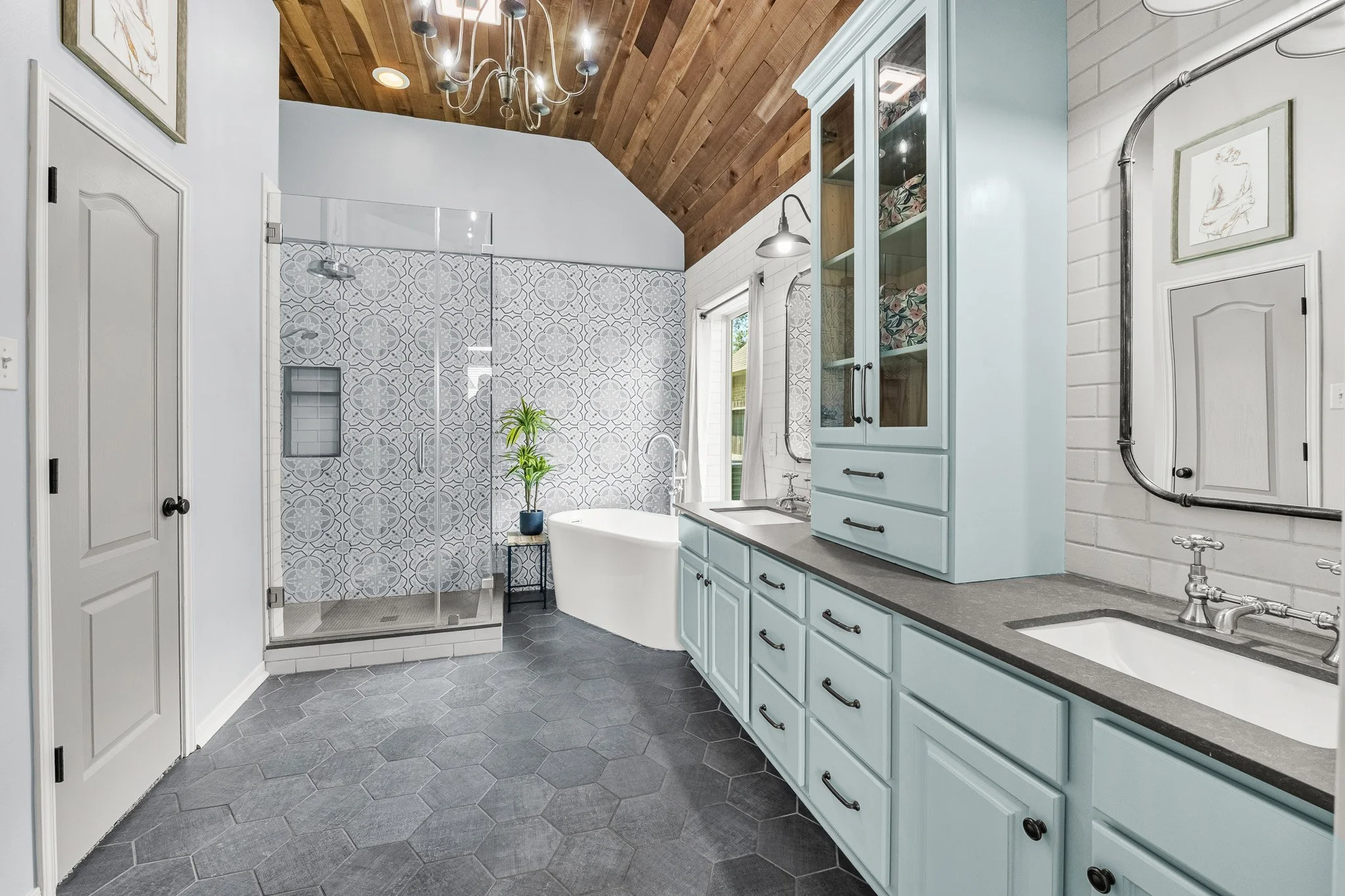 A bathroom with light blue cabinets, a double sink, a corner shower with patterned tiles, a freestanding bathtub, and a wooden-paneled vaulted ceiling with a chandelier.