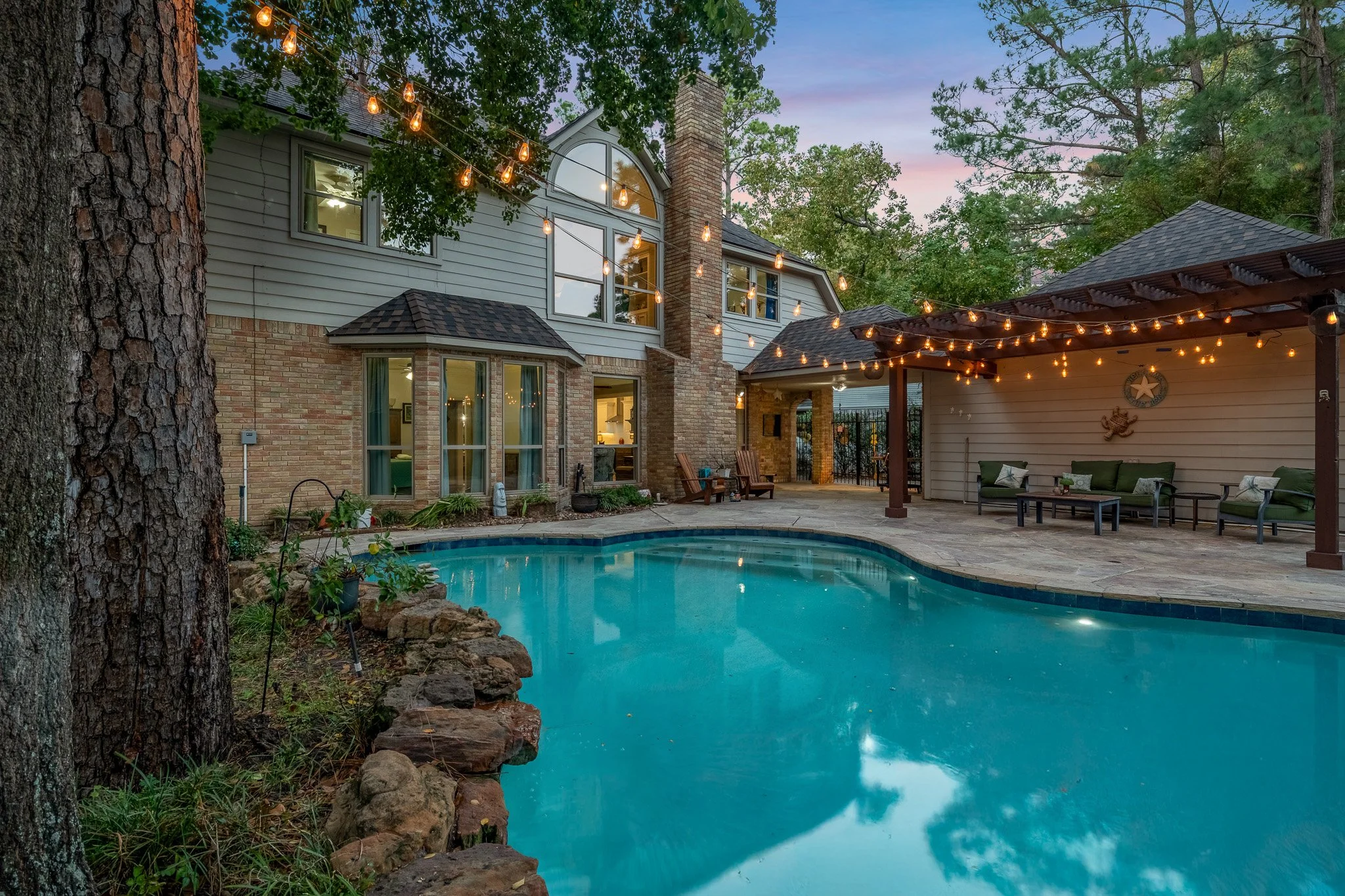 A backyard scene at dusk featuring a swimming pool, outdoor seating area with green couches under a pergola with string lights, a large two-story house with brick and siding exterior, and trees surrounding the area.