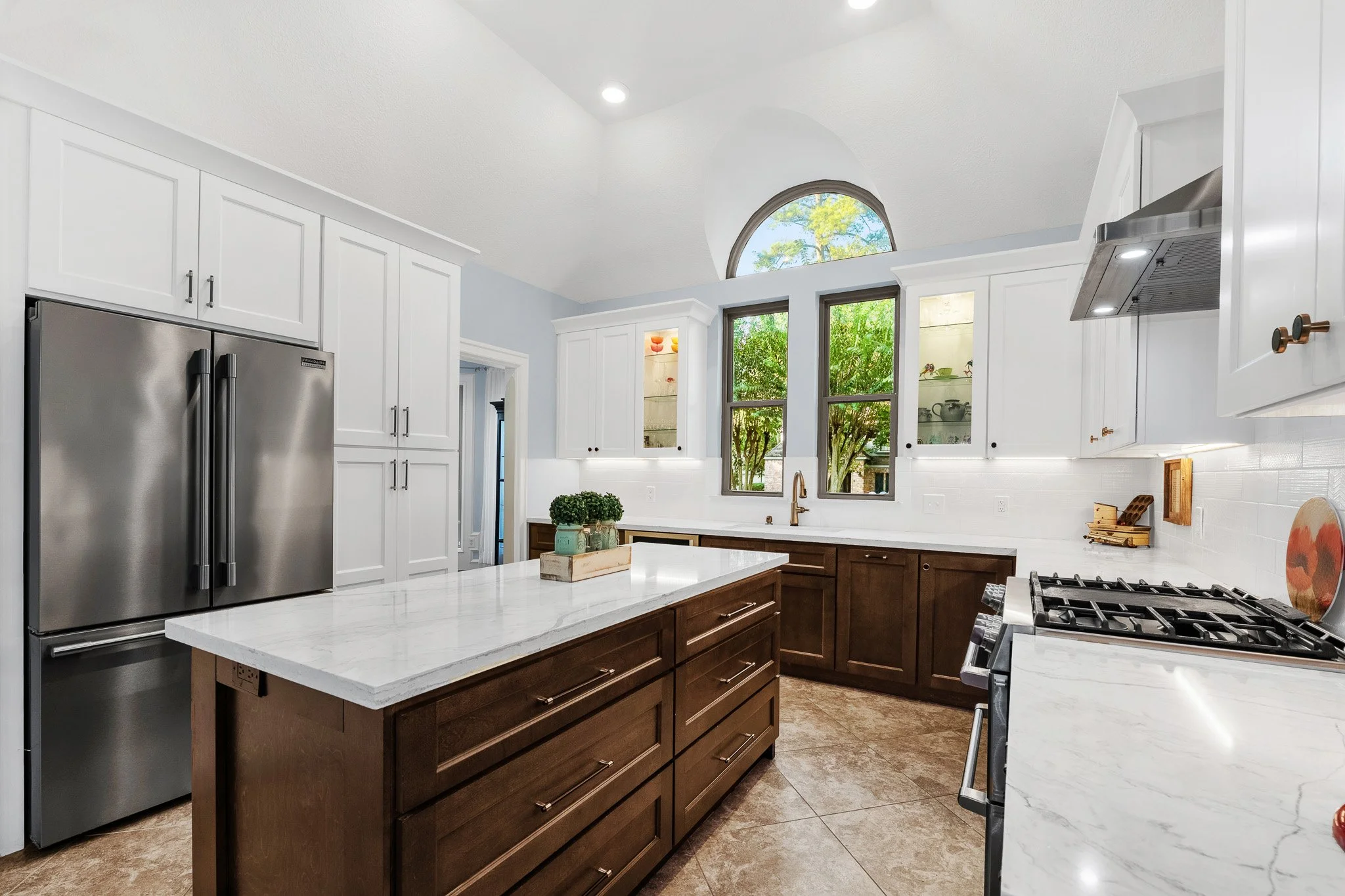 Modern kitchen with white upper cabinets, dark lower cabinets, marble countertops, large window, and stainless steel appliances.